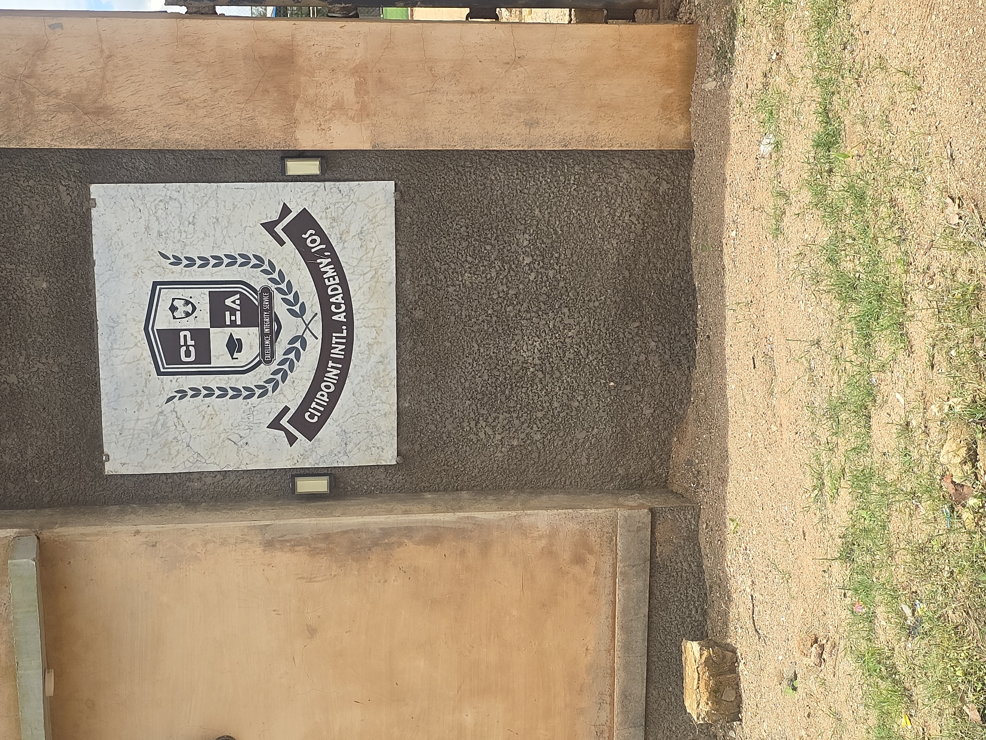 The image shows the entrance to Citipoint International Academy in Jos, Nigeria. The academy's crest is displayed prominently on a marble plaque mounted on a textured, dark grey wall. The crest features a shield with the letters "CP" and "EA", a graduation cap, and a laurel wreath. Below the crest, the academy's motto, "EXCELLENCE, INTEGRITY, SERVICE", is visible, along with the name "CITIPOINT INTL. ACADEMY, JOS". The wall is flanked by a lighter, textured beige wall. The ground in front of the entrance is sandy with patches of sparse green grass. A small, rough-hewn stone sits on the sandy ground to the left. The overall scene depicts the exterior of an educational institution.