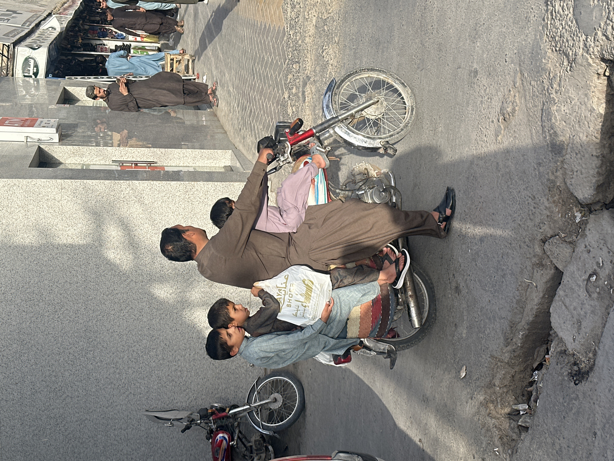 The photograph captures a man and three children on a motorcycle in an urban environment, likely within Quetta City Tehsil, Pakistan. The man, dressed in traditional Pakistani clothing, is operating the motorcycle. The children are seated behind him, one in the middle, and two behind him on the motorcycle seat. One of the children is holding a white plastic shopping bag.
The surrounding environment features paved streets with adjacent buildings, sidewalks, and other motorcycles parked nearby. A group of people are standing in front of the buildings, and the street exhibits signs of wear and tear.