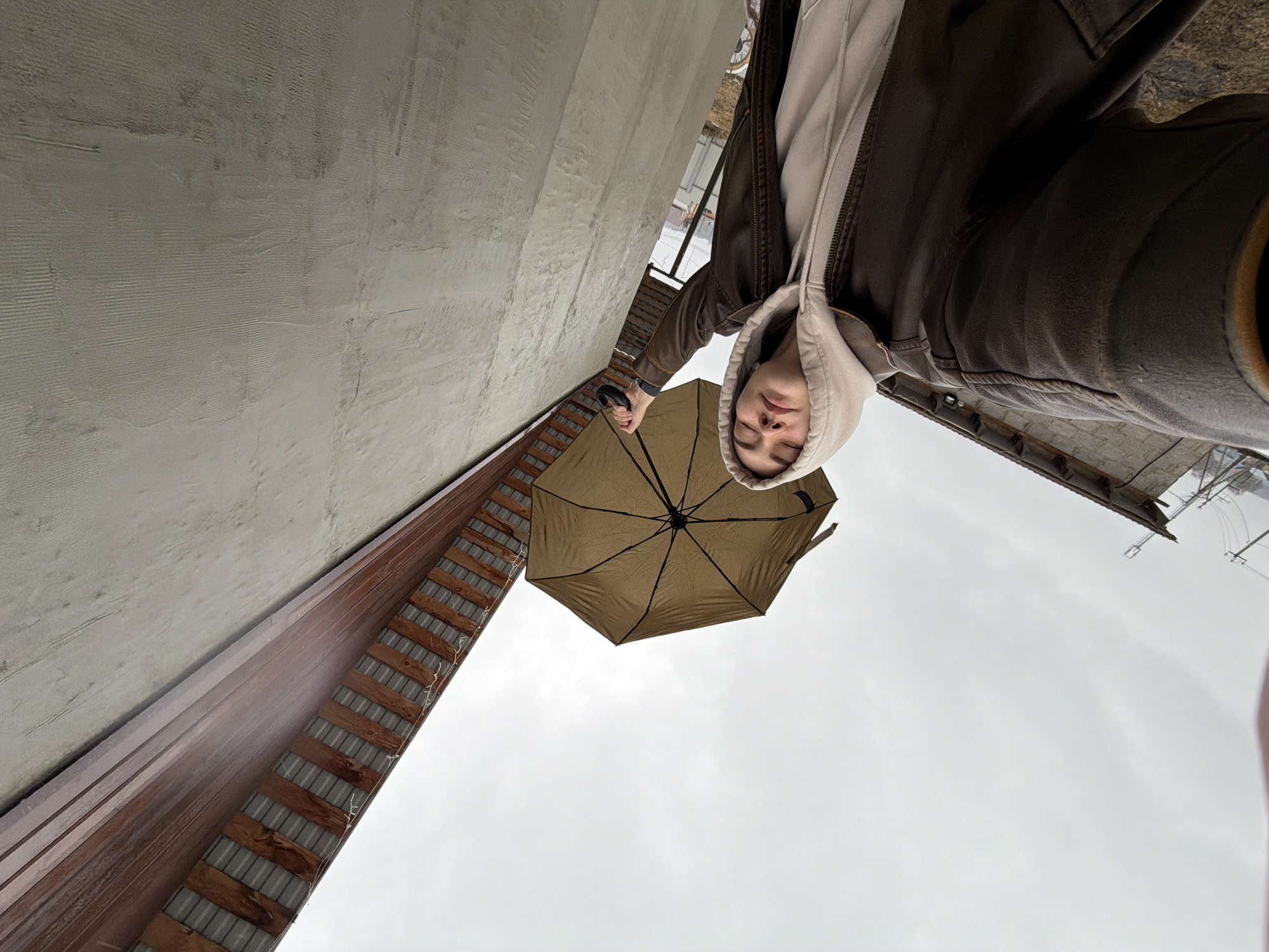 A person with a tan umbrella is photographed from a low angle near what appears to be a building. The person is wearing a brown jacket over a light-colored hooded sweatshirt. The umbrella is directly above the person. The sky is overcast and gray. Part of a building with horizontal boards is visible, as well as a concrete wall. The photo may have been taken in Chaikovsky, Russia.
