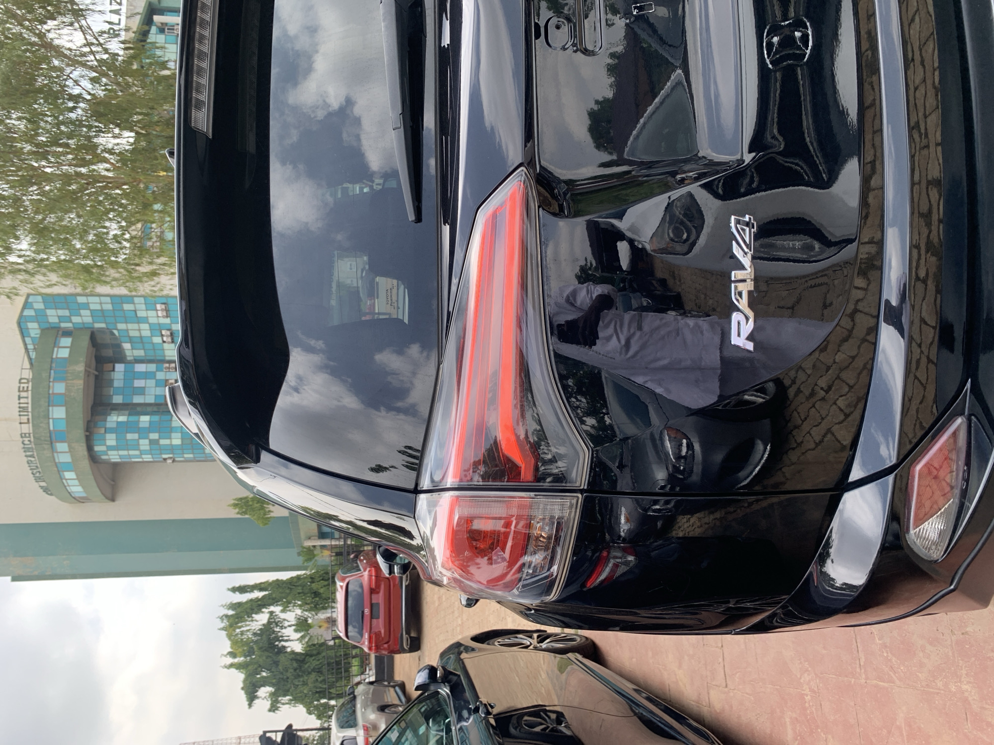 The image displays the rear and left side of a glossy black Toyota RAV4 SUV, parked outdoors under a cloudy daytime sky. The vehicle's 'RAV4' emblem and the Toyota logo are clearly visible on the tailgate. Its distinctive red and clear taillights are prominent, along with a rear spoiler that incorporates a brake light above the back window. The car's polished surface reflects the sky, clouds, and surrounding elements, including a person's reflection near the taillight, suggesting the photographer's presence.
Other vehicles, including a red SUV and a grey car, are partially visible, parked in the vicinity. The setting is an urban environment, likely a commercial parking area or street in Abuja, Nigeria. In the background, a multi-story building with blue-tinted windows and a modern facade is visible. Text on the side of this building reads "UNITYKAPITAL ASSURANCE LIMITED" and "UNITYKAPITAL PLAZA," providing a significant location cue. Trees and other green foliage dot the landscape around the buildings and parked cars. No specific activity is depicted other than the cars being stationary. The image is taken from a low angle, looking upwards towards the vehicle and the background.