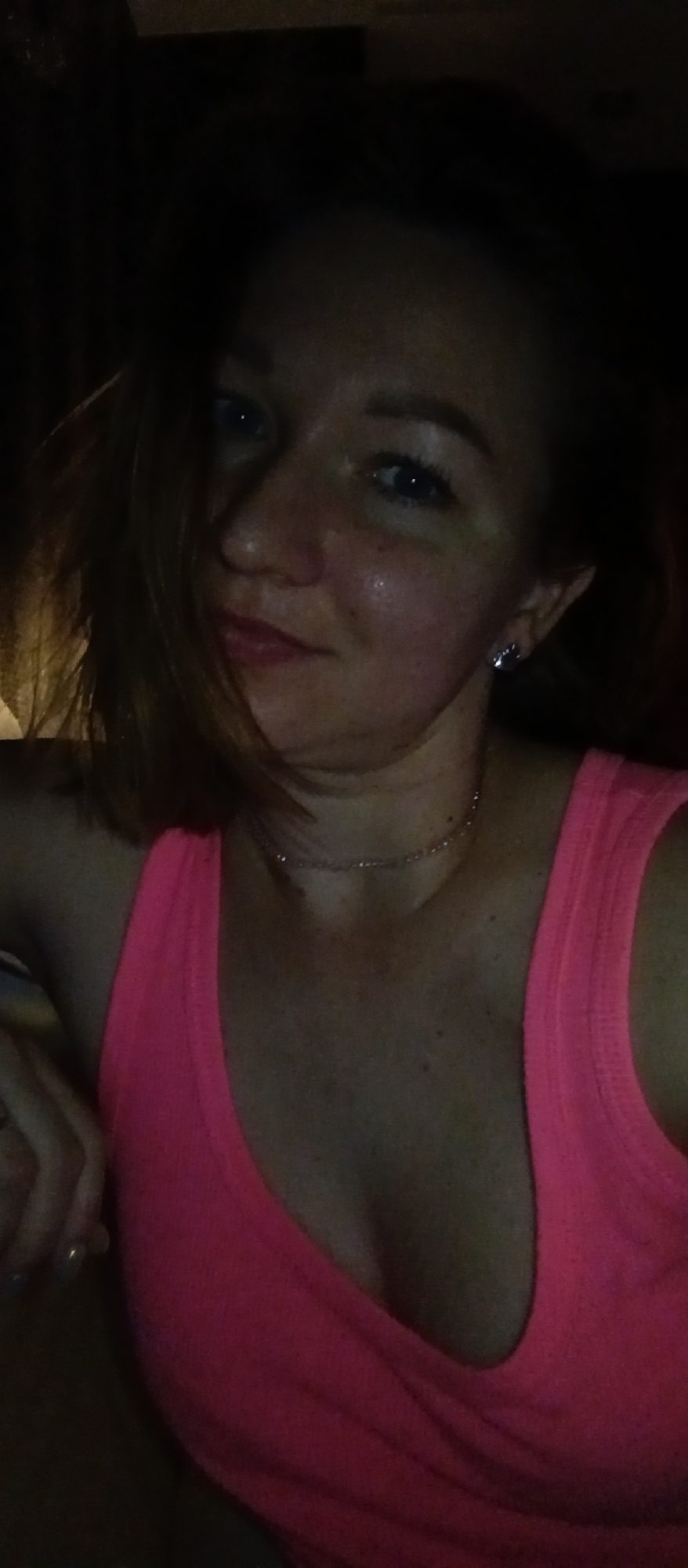 A woman with fair skin, blue eyes, and light red hair is pictured in a selfie. She is wearing a bright pink tank top and a delicate silver necklace. Her left hand rests on a surface outside the frame. The background is dark, suggesting an indoor setting. The lighting is dim.