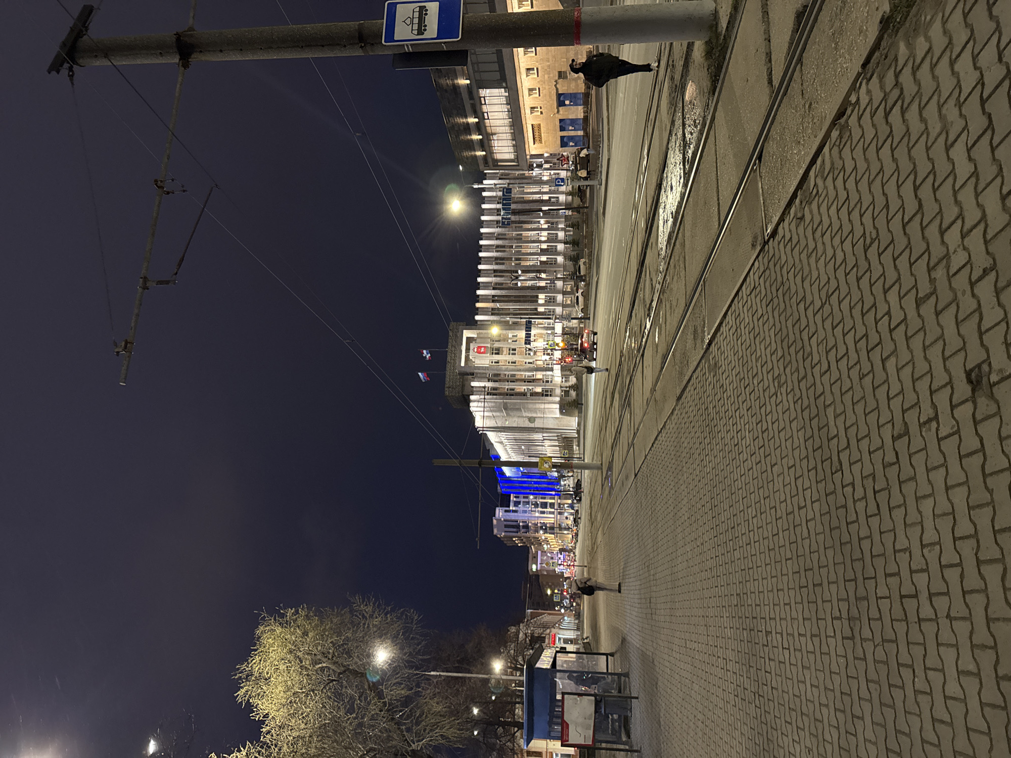 The image captures a night scene in an urban area, rotated 90 degrees counter-clockwise. Re-orienting it, the view is down a city street lined with buildings and a sidewalk.
In the foreground, a textured paved sidewalk, likely made of interlocking concrete blocks, stretches along the right side of the frame. Parallel to the sidewalk are tram tracks. A bare, deciduous tree, illuminated by a nearby streetlight, stands on the left. Further along the left side, a blue-framed bus stop or shelter is visible.
The street itself features multiple tram tracks and overhead electrical lines for public transport, suspended from poles that also hold streetlights. Buildings of various architectural styles line both sides of the street, most of them illuminated, indicating commercial or residential activity. Some buildings display many windows, while others have more solid facades. A prominent blue road sign with a white tram icon (indicating a tram stop) is attached to one of the overhead line poles. Another sign, a white "P" with an arrow, suggests parking.
Several figures are visible on the street and sidewalks, including a person standing near the tram tracks on the right and another further down the street on the left. In the distance, red traffic lights can be discerned. On poles near one of the buildings on the left, flags with horizontal white, blue, and red stripes (Russian flags) are flying.
The time of day is clearly night, with the sky a deep dark blue. Artificial lights from buildings, streetlights, and traffic signals provide the illumination. The street surface appears somewhat reflective in places, possibly from recent dampness or just the effect of the lights. The scene appears calm and quiet, characteristic of an evening in a city. This location is Perm, Russia.