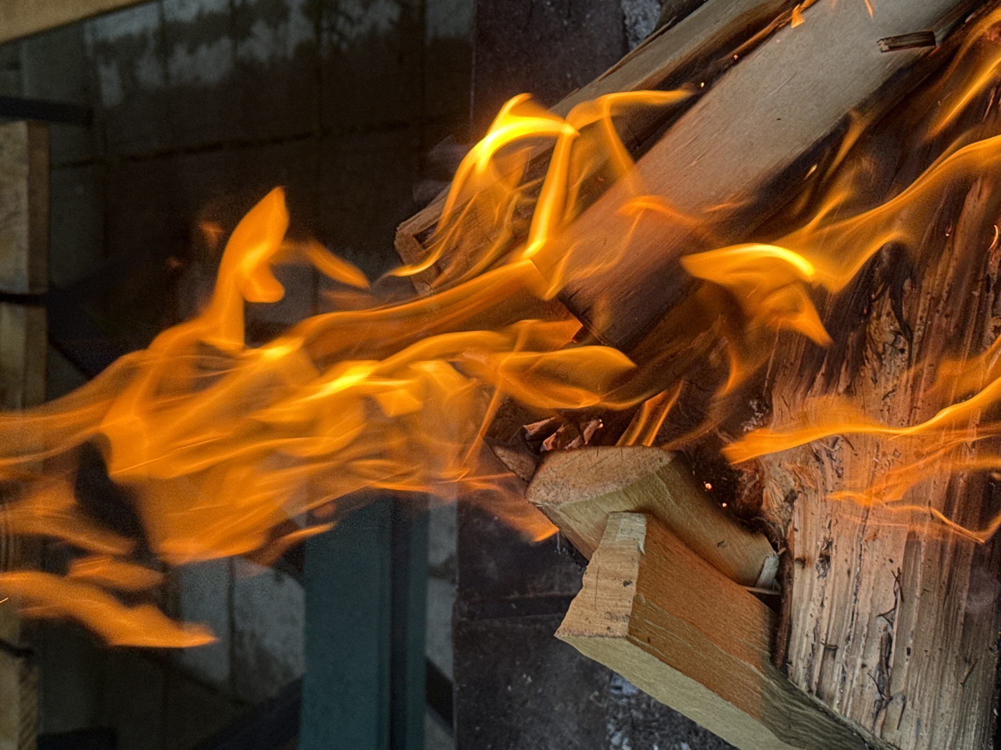The image shows a close-up view of a fire burning in an outdoor setting, likely a backyard or patio area. The flames are predominantly orange and yellow, with some hints of red, and they are actively consuming pieces of firewood. The firewood appears to be dry and stacked in a manner that suggests it's part of a fire pit or outdoor fireplace. The background is dark and slightly out of focus, but it seems to show a paved surface, possibly a patio or walkway, and some dark green elements that could be furniture or landscaping. There are no people visible in the image. The scene is illuminated by the fire itself. The location is specified as "Автомобилист-1 Снт, Russia," which is a garden association or cottage settlement.
