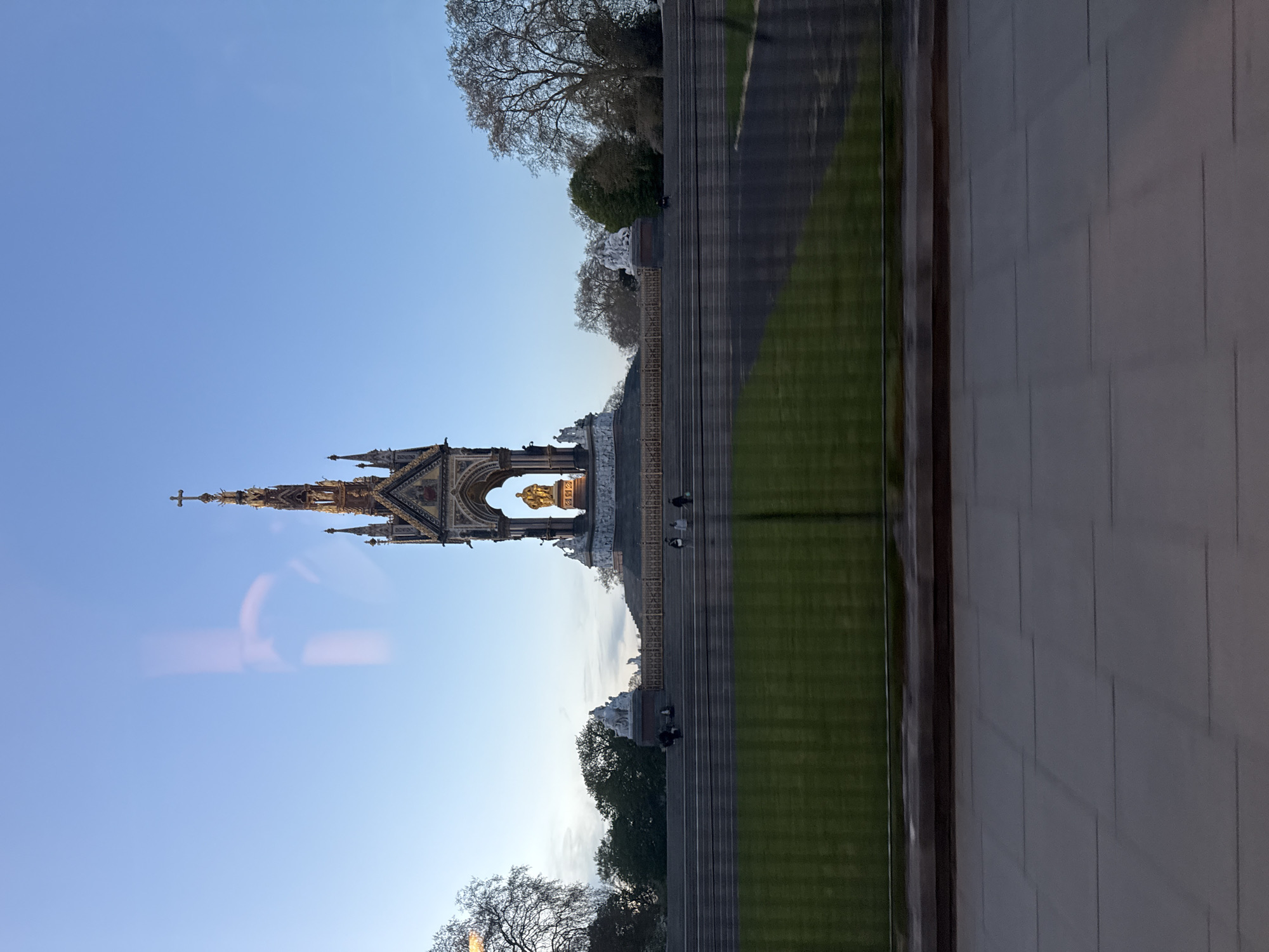The image shows the Albert Memorial in Kensington Gardens, London, UK. The monument is centered in the frame and is flanked by trees and a clear blue sky. In the midground, people sit on the steps leading to the monument. The image appears to be taken from inside a building looking out, as evidenced by the pane on the right and a possible reflection in the pane on the left.