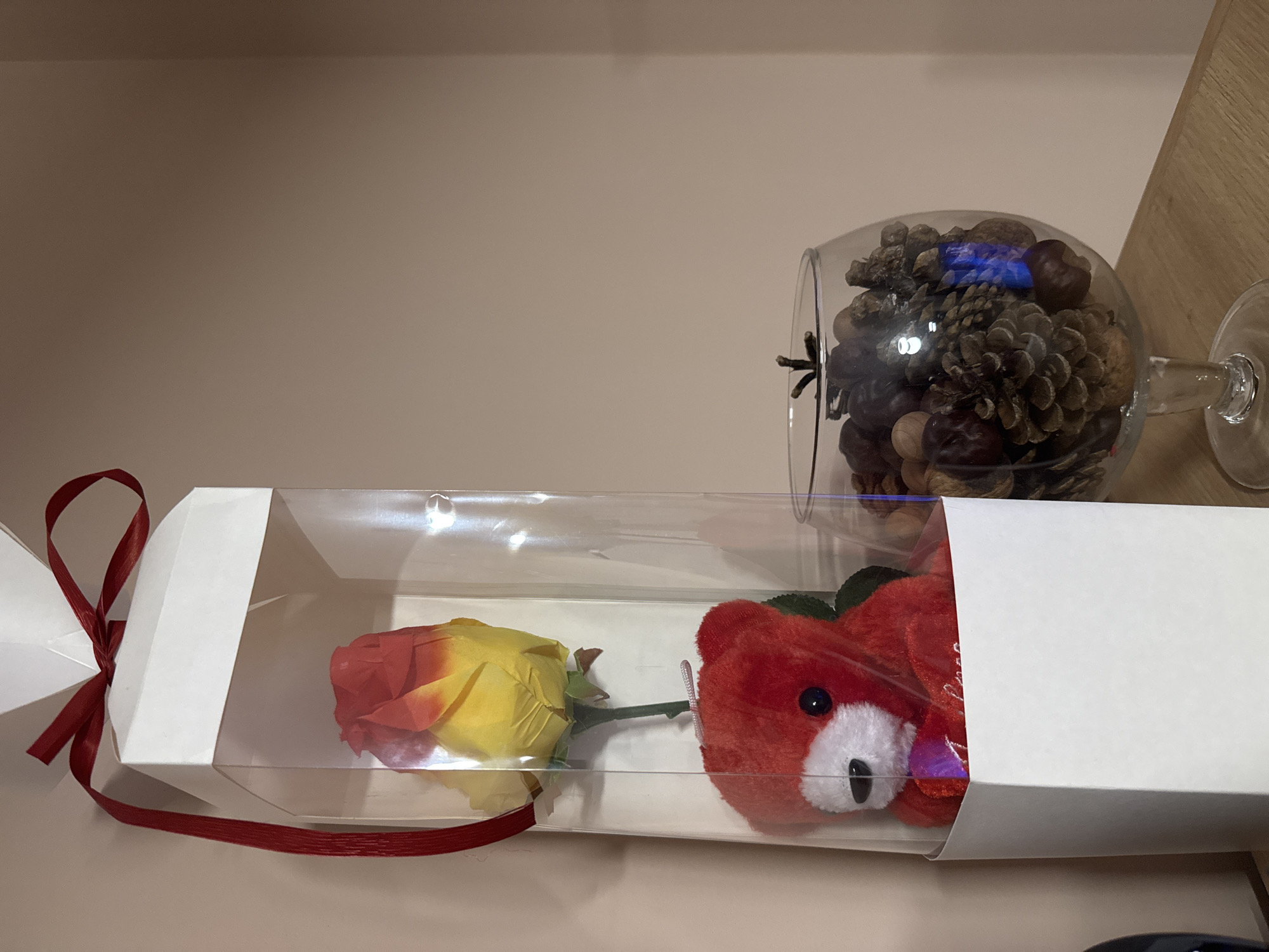 The image captures a static, indoor scene featuring a gift box and a decorative glass. In the foreground, a long, rectangular gift box with a transparent plastic top section lies horizontally. Inside the box, from left to right, is an artificial rose with petals transitioning from red to yellow, and a small, red plush bear with a white snout and black eyes. A red ribbon is tied around the left end of the gift box. To the right of the gift box, a clear glass goblet lies on its side, filled with an assortment of pinecones and chestnuts. A faint blue reflection or light is visible within the goblet. The background consists of a plain, light beige wall or surface, with a hint of a light wooden shelf or furniture piece in the upper right corner. No human activity is depicted. The lighting is soft and even, suggesting an indoor setting. There is no visible text.