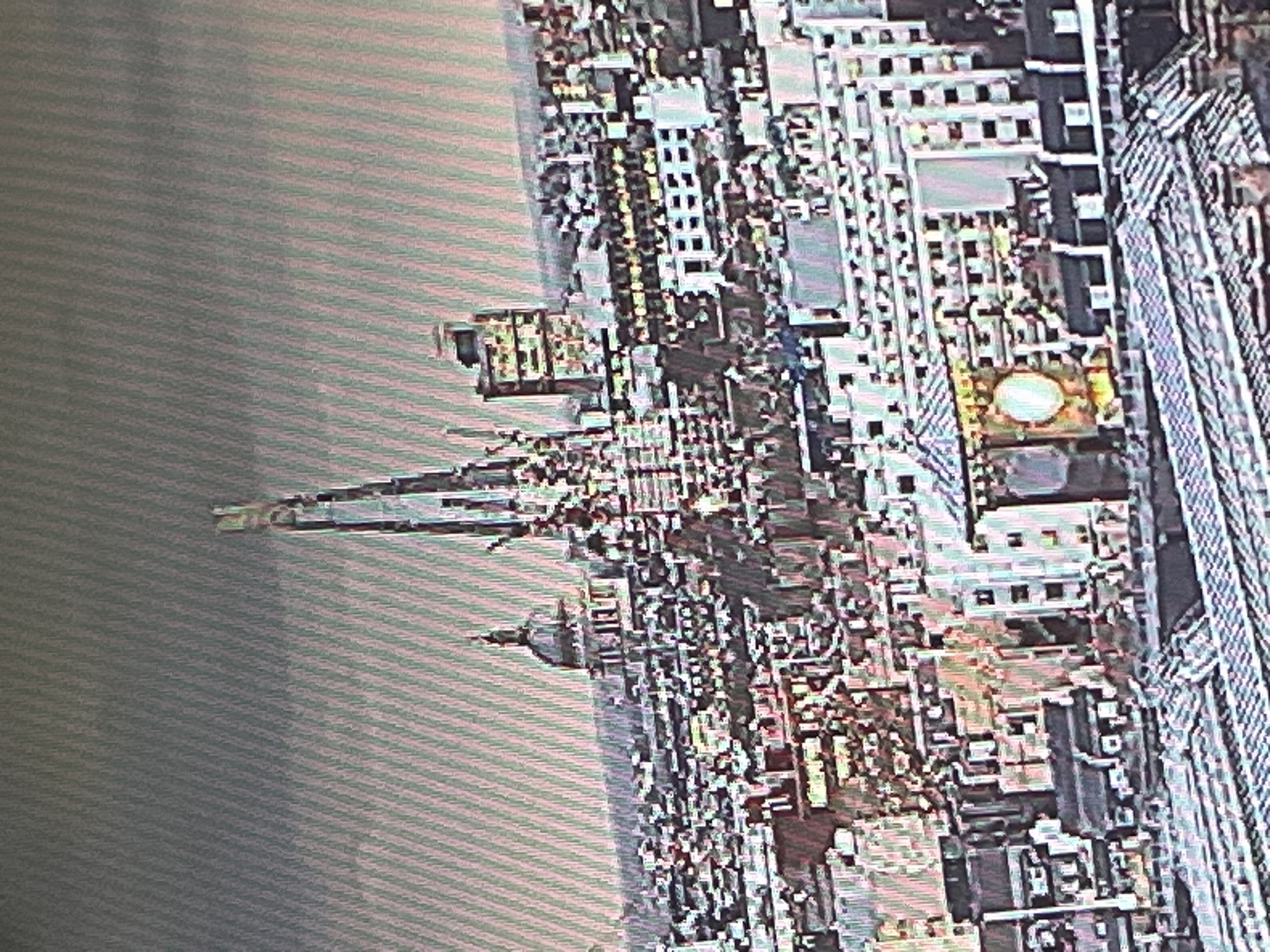 The image shows an aerial view of a city with numerous buildings, some of which are tall skyscrapers. The buildings exhibit a range of architectural styles, with a prominent dome-shaped structure visible on the left side of the frame, likely a church or cathedral. In the center of the image, two very tall, thin structures with intricate designs rise above the surrounding cityscape, appearing to be modern architectural marvels. The sky is overcast with a textured, striped pattern. The overall impression is of a densely populated urban environment, possibly a major metropolitan area. The image quality is degraded, with visible pixelation and a grainy texture, suggesting it may be a low-resolution photograph or a screenshot.