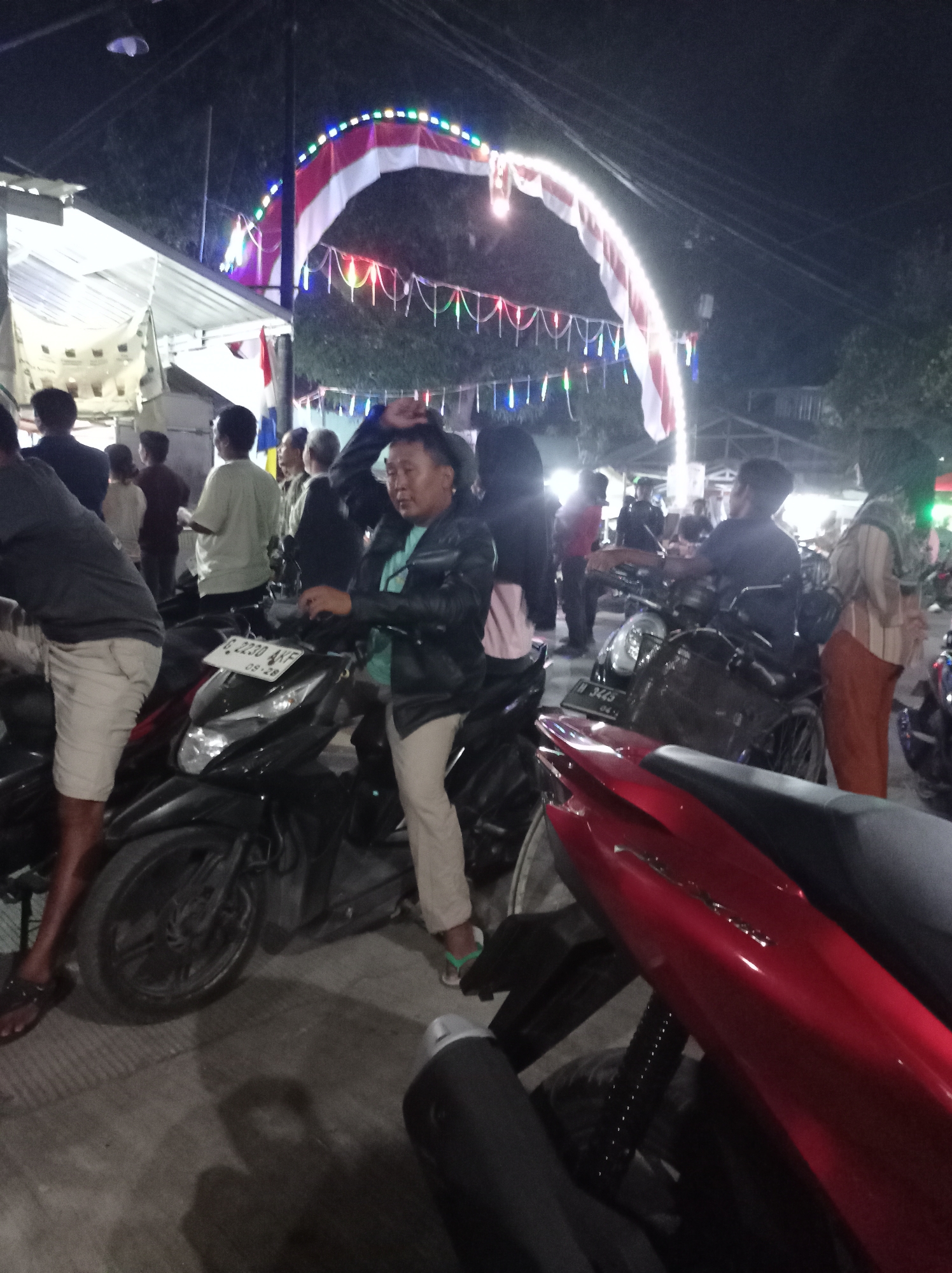 The image depicts an outdoor scene at night in Plumbungan, Indonesia, likely during a festive event. An arched decoration, featuring the Indonesian flag's colors of red and white, is illuminated by strings of colorful lights. Beneath this arch, a group of people are gathered around several motorcycles. One man, wearing a black jacket and light-colored pants, is standing next to a black motorcycle and appears to be interacting with it. Another person, wearing light-colored shorts, is also standing near a motorcycle. The ground is paved, and there are some structures and trees visible in the background. The overall atmosphere suggests a local gathering or celebration.