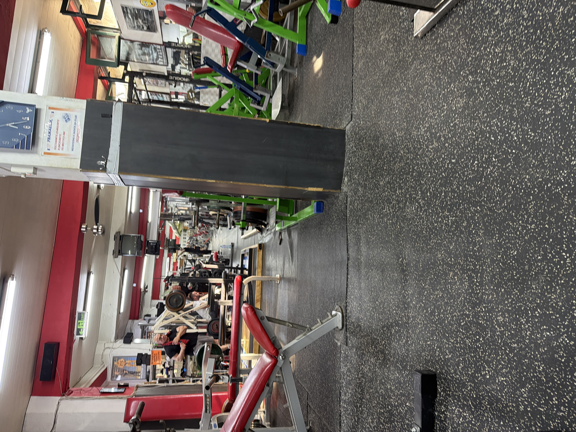 This image captures the interior of a gym or fitness center located in Plovdiv, Bulgaria. The scene is illuminated by overhead fluorescent lighting, revealing a spacious facility with a dark gray, speckled rubberized floor. Red structural beams and white walls form the upper perimeter of the room.
In the left midground, a bald, middle-aged man wearing a black t-shirt and gray shorts stands, a red towel draped around his neck. He is positioned near a red and silver adjustable weight bench with "MUSCLE T" branding visible. A barbell with weight plates rests on the floor nearby.
Towards the center-right midground, a prominent dark gray rectangular pillar rises from the floor to the ceiling. A blue-faced clock with white numbers is mounted near the top of the pillar, and below it, a white poster displaying Cyrillic text ("УВАЖАЕМИ" among other words) is affixed. A hook is taped to the pillar's side.
Beyond this pillar, in the midground and background, multiple individuals are engaged in physical activity. One man with dark hair, wearing a light-colored t-shirt, is seated on a weight bench beneath a barbell loaded with weight plates, suggesting a bench press exercise. An older man with white hair is partially visible near him, observing. Further back, various gym equipment is present, including multiple weight benches (some red), leg press machines with bright green and blue frames, barbell racks (one marked "ROGUE"), and stacks of weight plates. One individual in the distant background appears to be performing an exercise on a vertical rack.
The walls feature framed pictures, possibly related to bodybuilding, and a black rectangular speaker or vent is mounted high on the left wall. An "EXIT" sign with both English and Cyrillic text is visible on the upper left wall. A small television screen is also mounted on a wall in the midground-right.