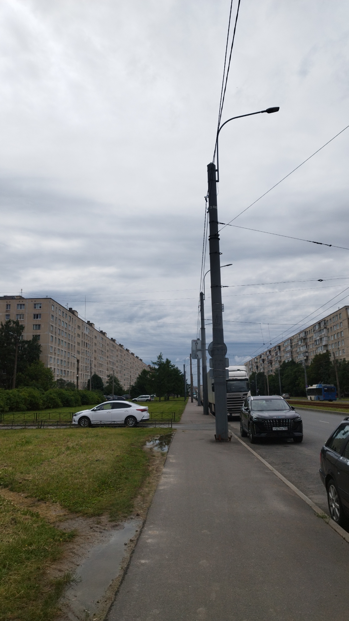 The image depicts a street scene in Saint Petersburg, Russia, under an overcast sky. A long, multi-story apartment building stretches along the left side of the street, with a grassy area and a fence separating it from the sidewalk. Several cars are parked or driving on the street. In the foreground, a sidewalk runs along the right side of the image. A large utility pole with streetlights and wires is prominently featured in the center right of the frame. Further down the street, there is a truck and more buildings. The overall impression is of a typical urban residential area on a cloudy day.