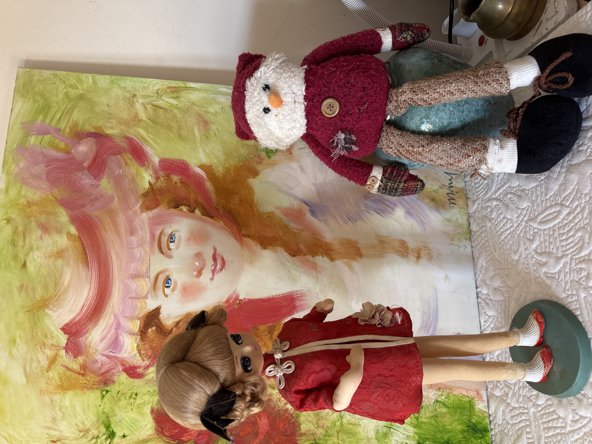 This image captures an indoor still life scene in Mirandela, Portugal. The primary subjects are two decorative figures positioned on a white, textured surface, possibly a quilted tablecloth, in front of a colorful painting.
The background features a vertical oil or acrylic painting on canvas. It depicts a stylized female bust with light blue eyes, pink cheeks, and closed lips, wearing a large, ornate pink and red hat or headpiece. The background of the painting is characterized by broad brushstrokes of green, yellow, and red. A partial signature, appearing as "miran," is visible in the lower right corner of the canvas.
To the left, a standing doll is presented. It possesses a light complexion, large dark blue eyes, and light brown hair styled in two prominent buns adorned with small black bows. The doll wears a long-sleeved, textured red dress with a white trim running down the front and white frog closures at the neckline. A light-colored floral embellishment is attached to the right side of its dress. The doll stands on an oval, teal-colored base, wearing white striped socks and red shoes with white detailing.
To the right, a plush, seated figure, resembling a snowman or festive character, rests on a glossy, teal-colored vase-like object. This figure has a white, textured head with black bead eyes and an orange, carrot-like nose. It wears a red, textured, long-sleeved shirt with a large wooden button centered on the chest and a small, frayed fabric patch to the left of the button. Its hands are covered by checkered red, black, and white mittens, each adorned with two small buttons. The figure's legs are made of light brown, textured fabric, and it wears white socks and black felt shoes with brown laces.
To the far right, partially visible behind the plush figure, are a white coiled tube and a small brass or copper jug. The background wall to the right of the painting is plain and light-colored.