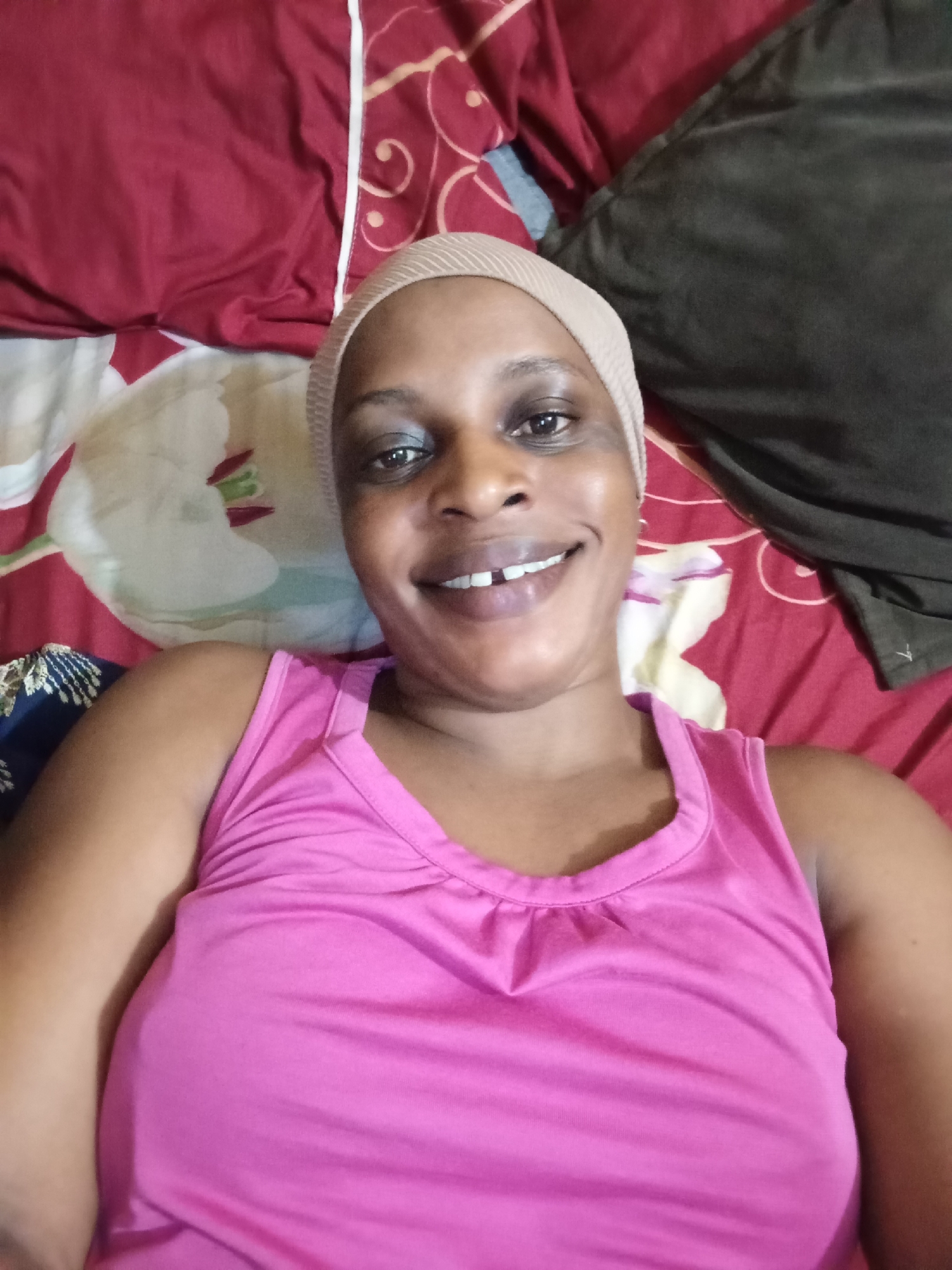 A medium shot depicts a Black woman lying on her back on a bed covered with a colorful floral-patterned bedspread. She is wearing a pink tank top and a beige head covering. She is smiling, revealing her teeth. The background includes a red sheet with a floral pattern and a dark-colored pillow or folded material. The photograph was taken in Narayi, Nigeria.