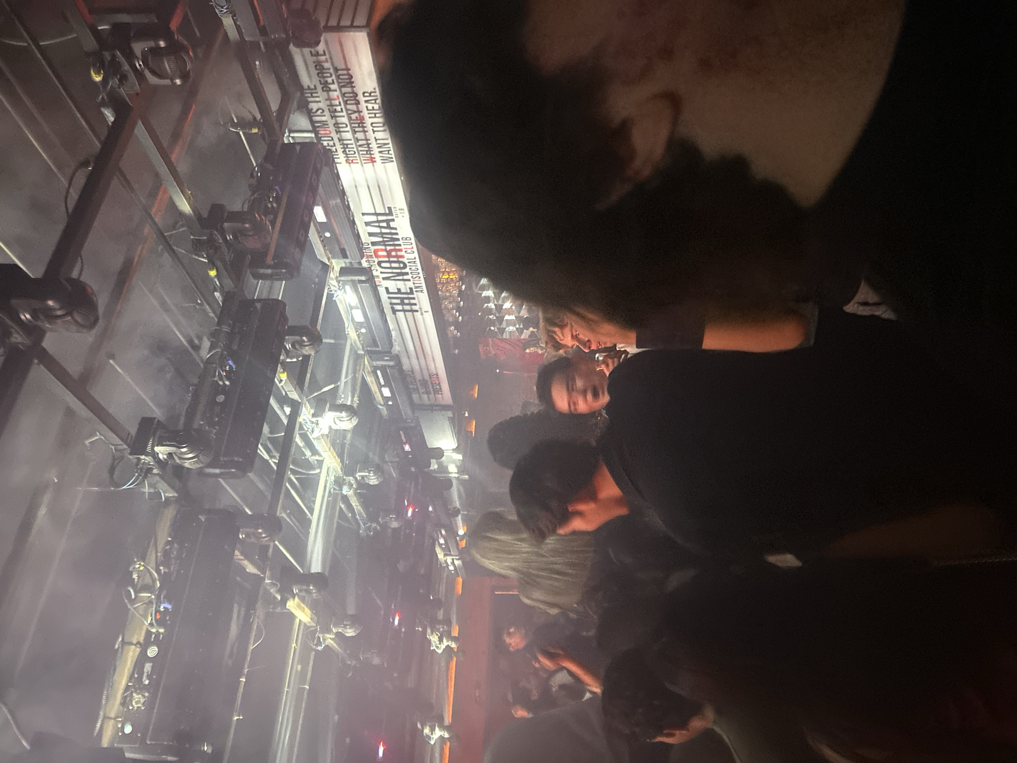 The image shows a crowded indoor space, possibly a club or theater in San Bernardino Tlaxcalancingo, Mexico. There is a banner or sign that reads "THE NORMAL ANTISOCIAL CLUB," with the tagline "Freedom is the right to tell people what they do not want to hear." The crowd includes a mix of people, with some faces visible and others obscured. Above the crowd are metal structures, possibly lighting rigs or stage equipment, with attached spotlights. The environment appears dimly lit with smoke or haze adding to the atmosphere.