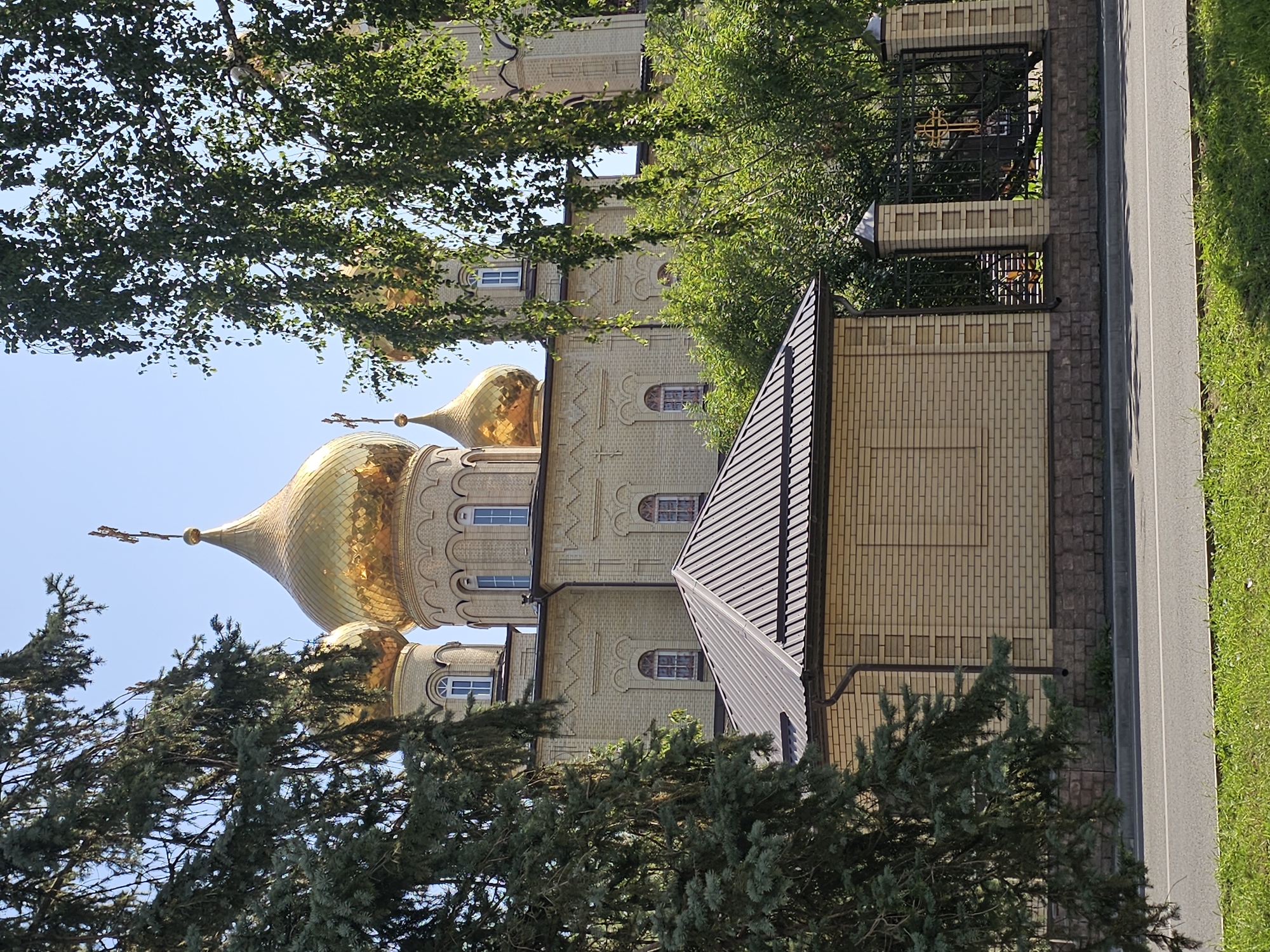 The image depicts the Orthodox Church of the Nativity of Christ in Cherkessk, Russia. The main facade of the church, constructed from light-colored brick, features multiple golden domes topped with crosses. A smaller, dark-roofed brick building is situated in the foreground, partially obscuring the church. Lush green trees, including a large fir tree on the left and deciduous trees with abundant foliage on the right, frame the view of the church. A black wrought-iron fence with decorative pillars and a golden cross emblem is visible on the right side, bordering the church property. The scene is set against a clear blue sky, and the foreground includes a paved road and a strip of green grass. No people are present in the image.