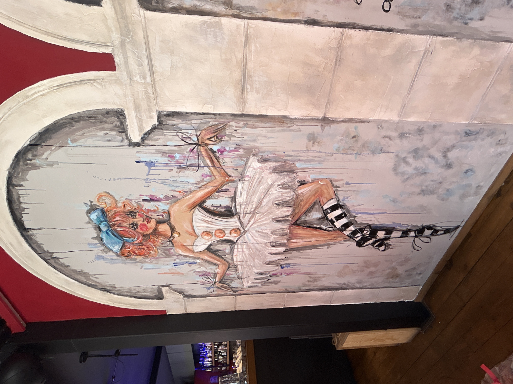 Ceiling mural in a Plovdiv, Bulgaria bar or restaurant, depicting a stylized female figure in a white tutu and striped stockings, likely a ballerina, set against a gray and white faux wood-plank background with dripping paint effects. The mural is framed by a red and white arched architectural detail. Below, a partial view of a bar with bottles is visible, and to the right, a wooden wall.