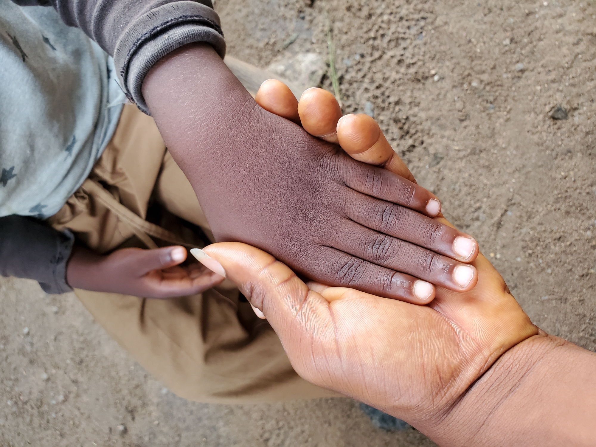 The image shows two hands clasped together. The hand on top is smaller and has a darker skin tone, belonging to a child wearing a grey shirt with star patterns and khaki pants. The hand underneath is larger with a lighter skin tone, likely an adult. The background appears to be an unpaved ground with dirt and small rocks. Given the prompt, the location is presumed to be Mararaba, Nigeria. The interaction depicted is one of holding hands.