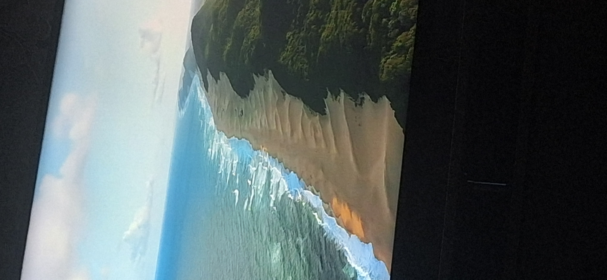 This is an aerial, high-angle, wide shot of a coastal landscape. The image features the ocean on the left, with waves gently breaking along a sandy beach. To the right of the beach are rolling hills covered in dense green vegetation. The sky above is a pale blue with scattered white clouds. The bottom portion of the image is dark, obscuring the ground and any potential human activity. The scene depicts a natural environment and does not provide any information about the city of Bila Tserkva, Ukraine.