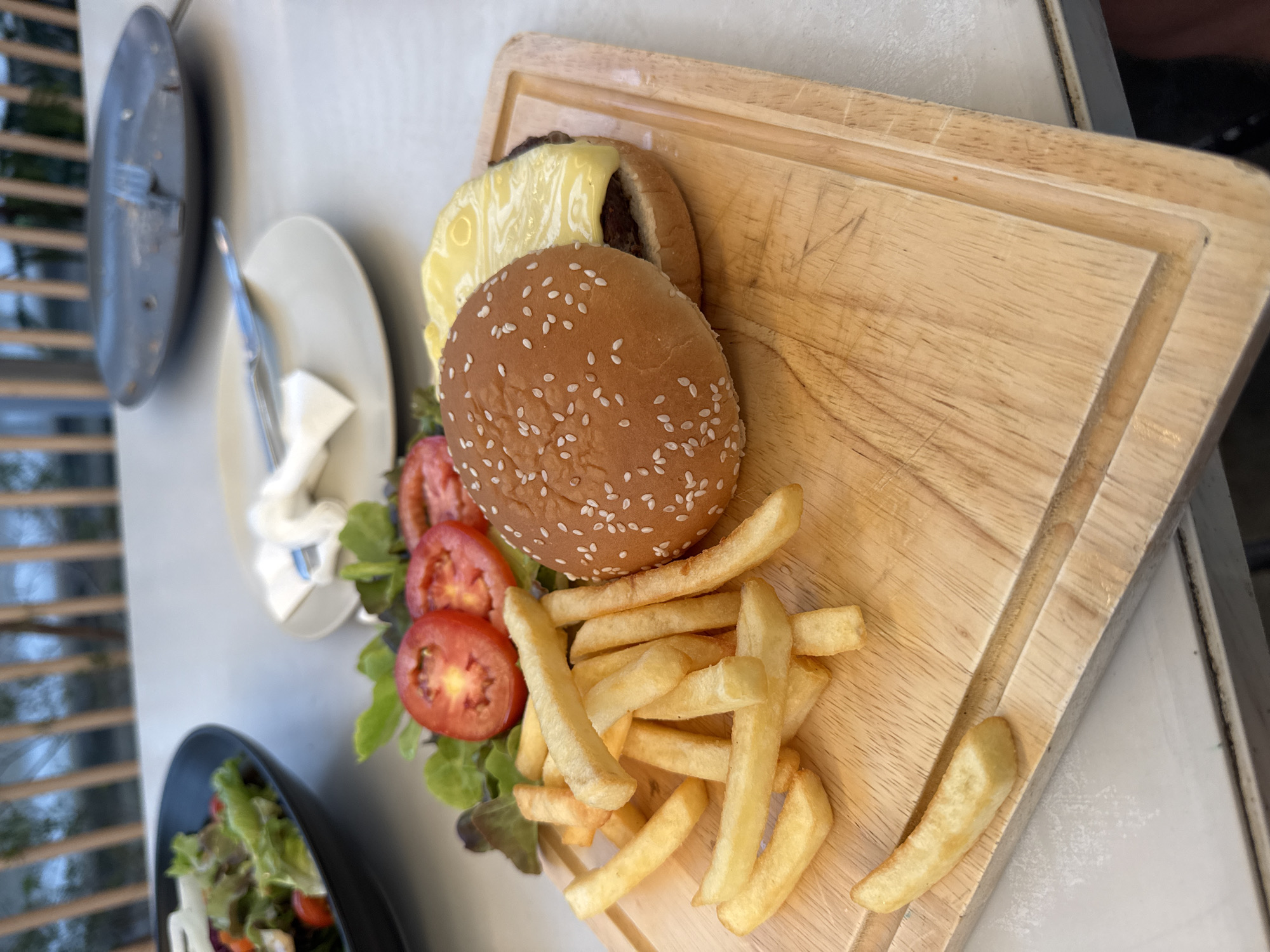 In a close-up, eye-level shot, a wooden cutting board holds a classic cheeseburger and a pile of golden-brown french fries. The burger, topped with melted yellow cheese, sits on its bun, which is sprinkled with sesame seeds. Sliced tomatoes and green lettuce leaves add a touch of color to the plate. The fries are scattered around the burger, some appearing crisp and others soft. The scene is set on a white table, with other plates and cutlery visible in the blurred background, suggesting a meal is being served outdoors or in a casual dining setting, possibly at a restaurant.