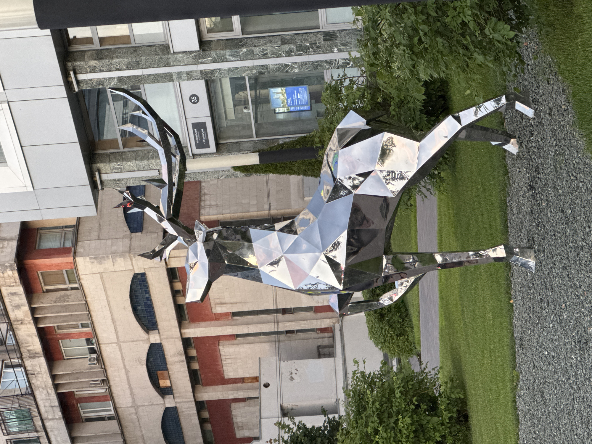 A chrome-plated, geometric sculpture of a deer stands on a patch of gravel and grass, in front of a building with a modern facade. The deer is rendered in a faceted style, with sharp angles and a highly reflective surface that mirrors its surroundings. Behind the sculpture, a multi-story building exhibits a Brutalist architectural style, characterized by its exposed concrete and red brick accents. The building features numerous windows, some arched and others rectangular, along with visible air conditioning units. To the right of the deer sculpture, a portion of a grey, stone-clad building is visible, with signage indicating street names in Russian: "ул. Октябрьская" and "ул. Варварская". A number "35" is also present on the building facade. The environment appears to be an urban setting, possibly a public space or courtyard, with lush green bushes and mowed lawn surrounding the sculpture. The lighting suggests it is daytime, likely late afternoon or early evening, given the warm tones of the light. No people are visible in the image.