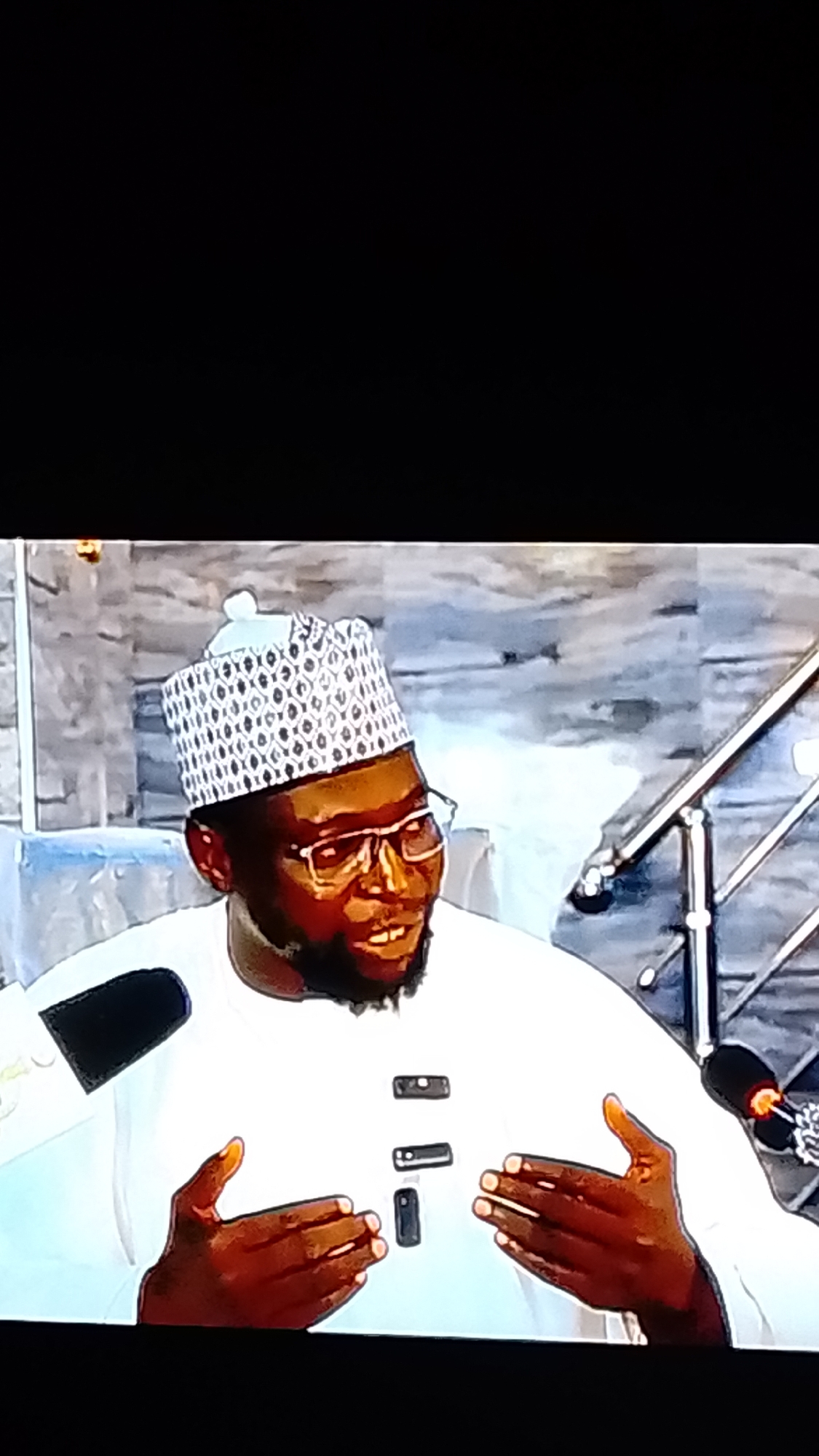 A medium shot, from a slightly low angle, shows a Black man wearing a white tunic and a patterned Kufi hat, speaking into a microphone. He wears glasses and has a beard. His hands are raised in front of him. The background consists of a textured wall and a metal railing. The lighting is dim.