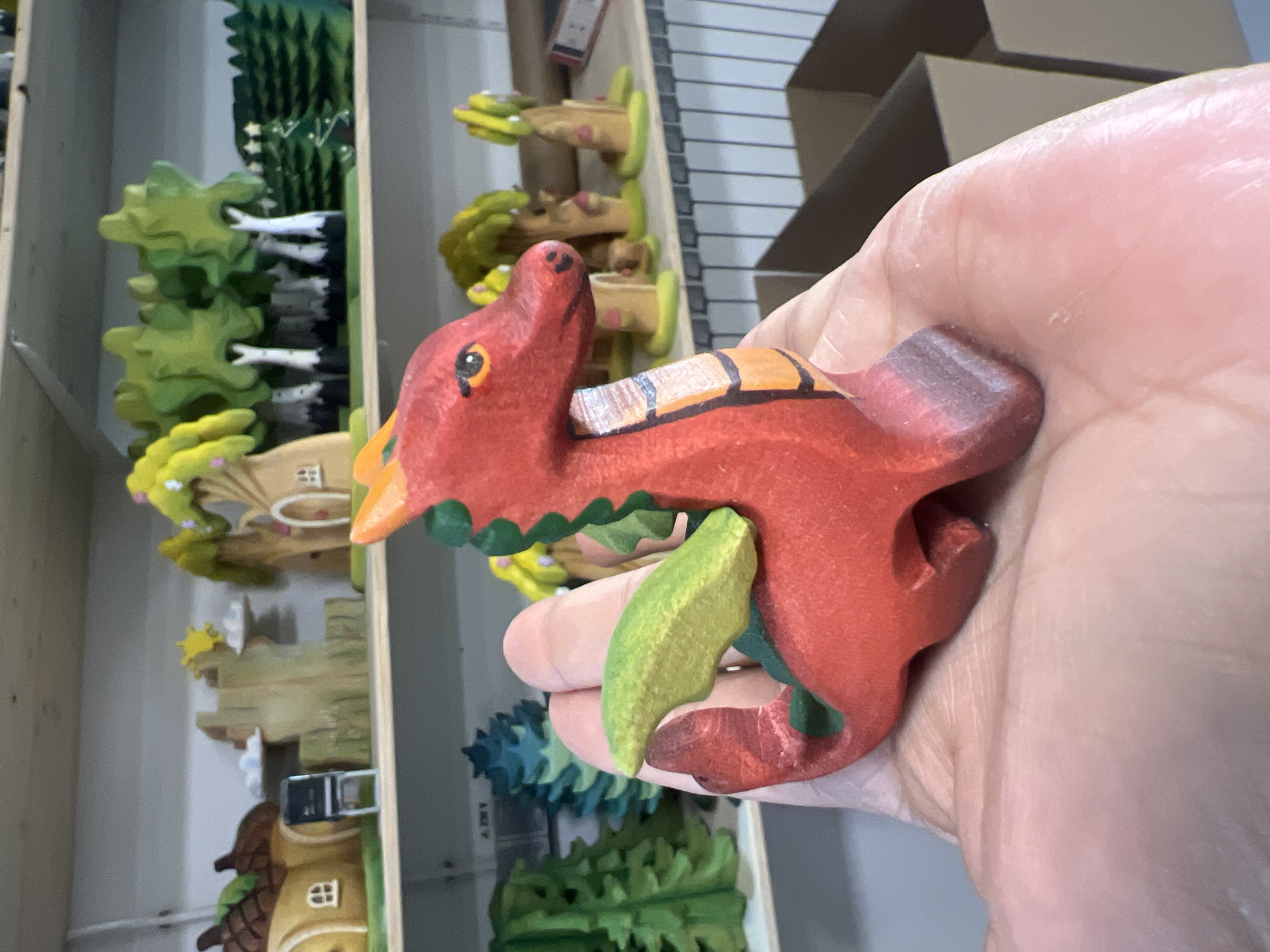 The image shows a person's hand holding a red wooden toy dragon. The dragon has a green leaf-like object in its mouth and orange and black stripes on its back. The background is blurred, but appears to be a shelf filled with more wooden toys, including trees and houses. The lighting suggests an indoor setting, possibly a shop or a home. The focus is on the toy dragon, held up by the person's hand. There is no discernible text in the image.