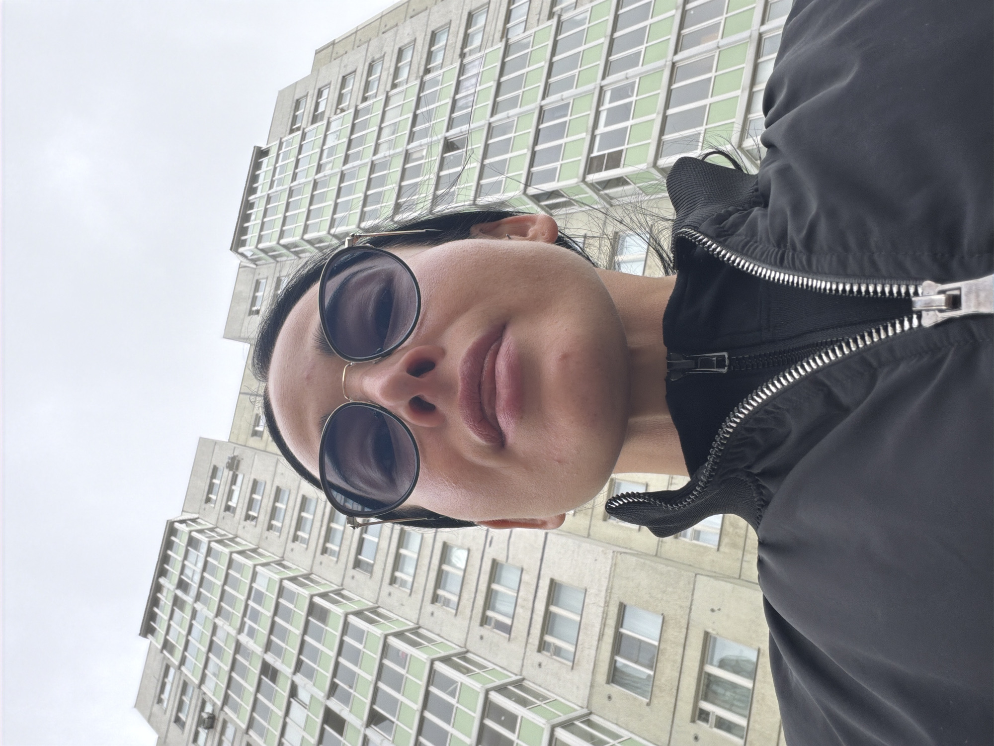 An upward-angled selfie captures a person against the backdrop of a tall, light-colored building in Saint Petersburg, Russia. The individual, seen from the nose up, wears large, round, dark sunglasses with thin frames, obscuring their eyes. Their lips are slightly parted, and their chin is visible. They are wearing a black jacket with a prominent silver zipper, partially unzipped. The person's dark hair is visible around their face.
The building in the background features a distinctive architectural style, with many rows of rectangular windows. Some window sections have a light green or greenish-white tint, forming a grid-like pattern across the facade. The sky above is overcast and a uniform light grey, indicating a cloudy day. The overall impression is a casual, low-angle portrait set against urban architecture. There is no visible text in the image.