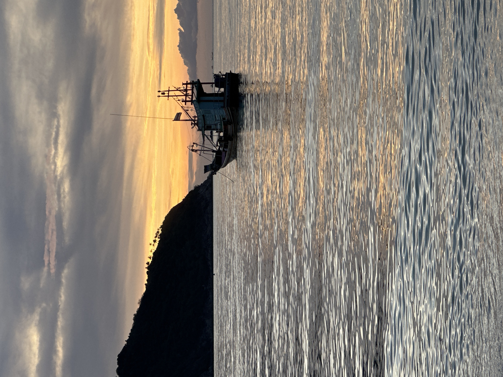 The image shows a fishing boat anchored off the coast of Baan Chaloklum, Thailand at dawn or dusk. The boat is silhouetted against the horizon and sits on calm water. The sky is an interplay of grey clouds, yellow and orange glow of the sun. The land is shown on the left side of the frame.