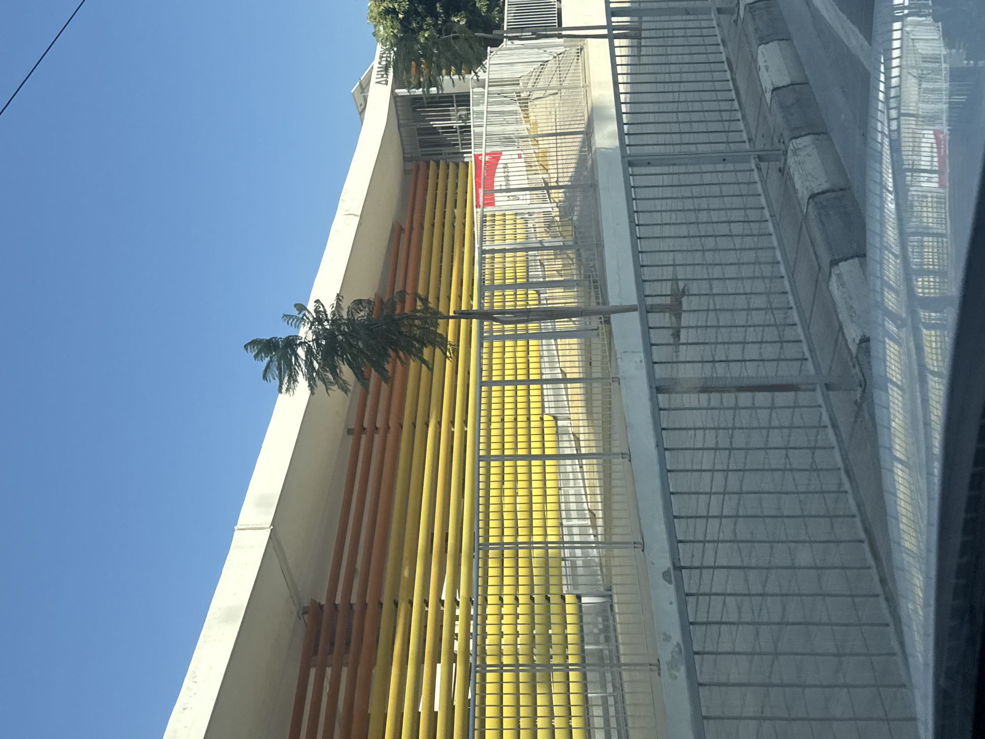 The image captures the exterior of a building in Mesa Geitonia, Cyprus. The building features a white overhang, beneath which are horizontal slats in alternating shades of orange and yellow. A metal fence runs along the front of the building, enclosing a sloped walkway leading to an entrance. Beyond the fence, the building's facade is visible with more yellow slats and a red and white sign displayed near the entrance. The scene is set against a clear blue sky, and a thin, sparse tree stands in front of the building, its branches swaying slightly. The foreground shows a reflection of the building and sky in what appears to be a vehicle's surface, along with a section of pavement and a curb. There are no people visible in the image.