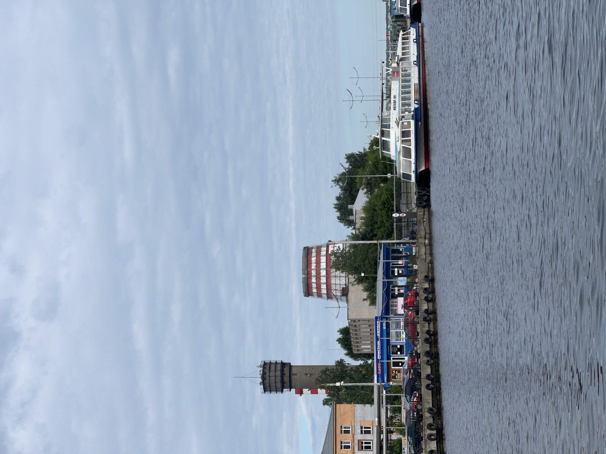 The image presents a waterfront scene in Izhevsk, Russia, under an overcast sky. A dark body of water occupies the foreground, exhibiting ripples on its surface. A concrete pier runs horizontally across the mid-ground, featuring numerous black rubber tires serving as fenders. Several cars, including black and red sedans, are parked on the pier behind the fenders. On the pier, a blue building with multiple windows and doors displays text identifying it as "КАФЕ - ПРИСТАНЬ - ИЖЕВСК" (Cafe - Pier - Izhevsk).
To the right of the blue building, a white and blue passenger boat, clearly labeled "МОСКВА-101" (Moskva-101), is docked at the pier. Part of another boat is visible docked immediately to its right.
In the background, various structures rise above the pier. On the far left, a multi-story building with yellow and orange facade and white windows is visible. To its right, a tall, grey concrete tower with an antenna-like structure on its top stands prominently. Behind and to the right of this tower, a slender red and white striped chimney is partially obscured. Further to the right, a large, hyperboloid cooling tower, also striped in red and white, dominates the skyline. Additional grey industrial-style buildings and green trees are visible between and around these larger structures. No individuals are discernible in the scene.