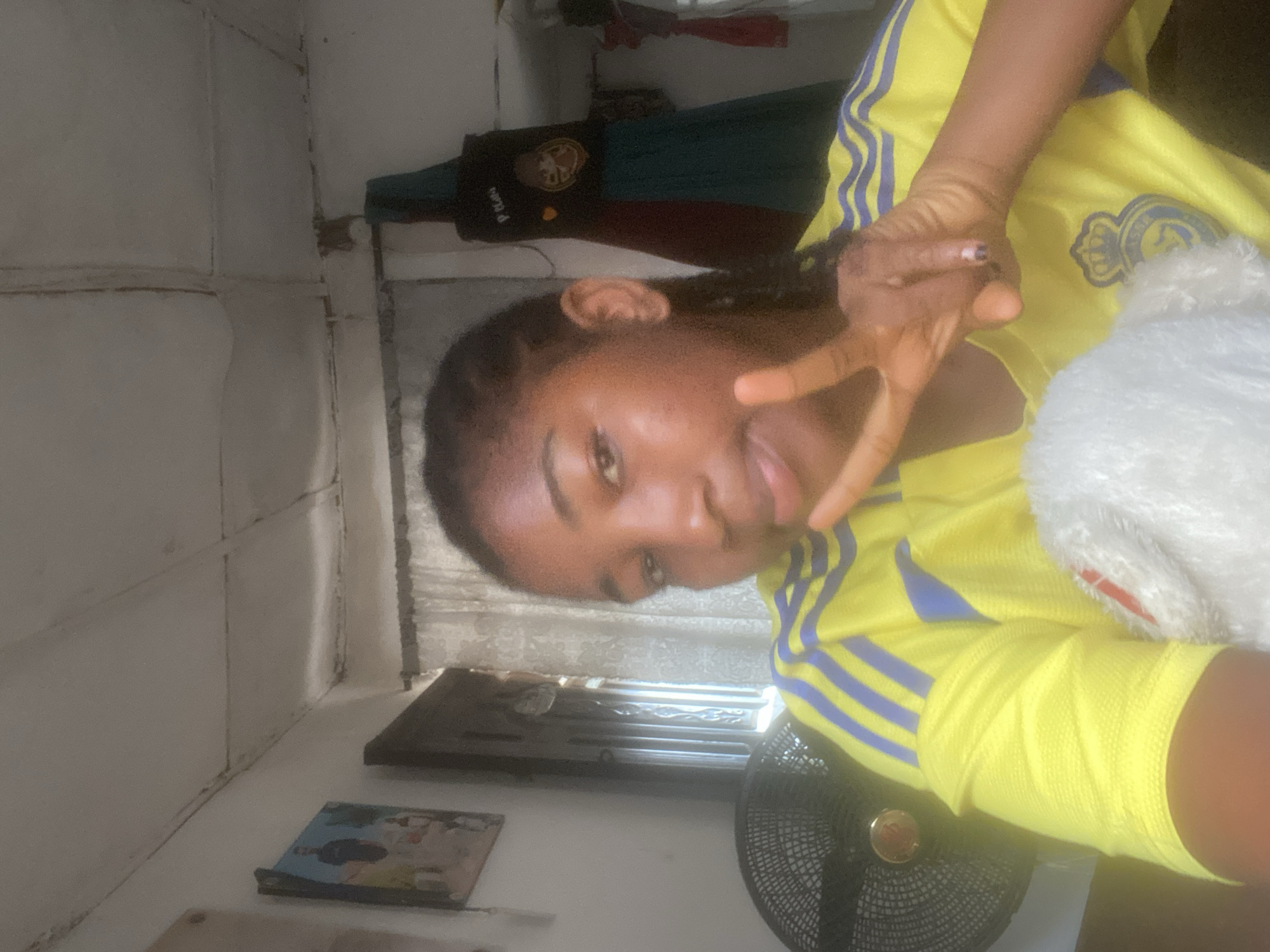 A young woman is posing indoors, making a peace sign with her right hand. She's wearing a yellow and blue athletic jersey. Her left arm is wrapped with a yellow bandage. Beside her is a white plush object. In the background is a bedroom setting, including an electric fan, framed pictures on a white shelf, and a closed window. The location is likely in a residential area, and given the prompt, it is inferred that it is in Lokoja, Nigeria. A black t-shirt hangs on the wall.