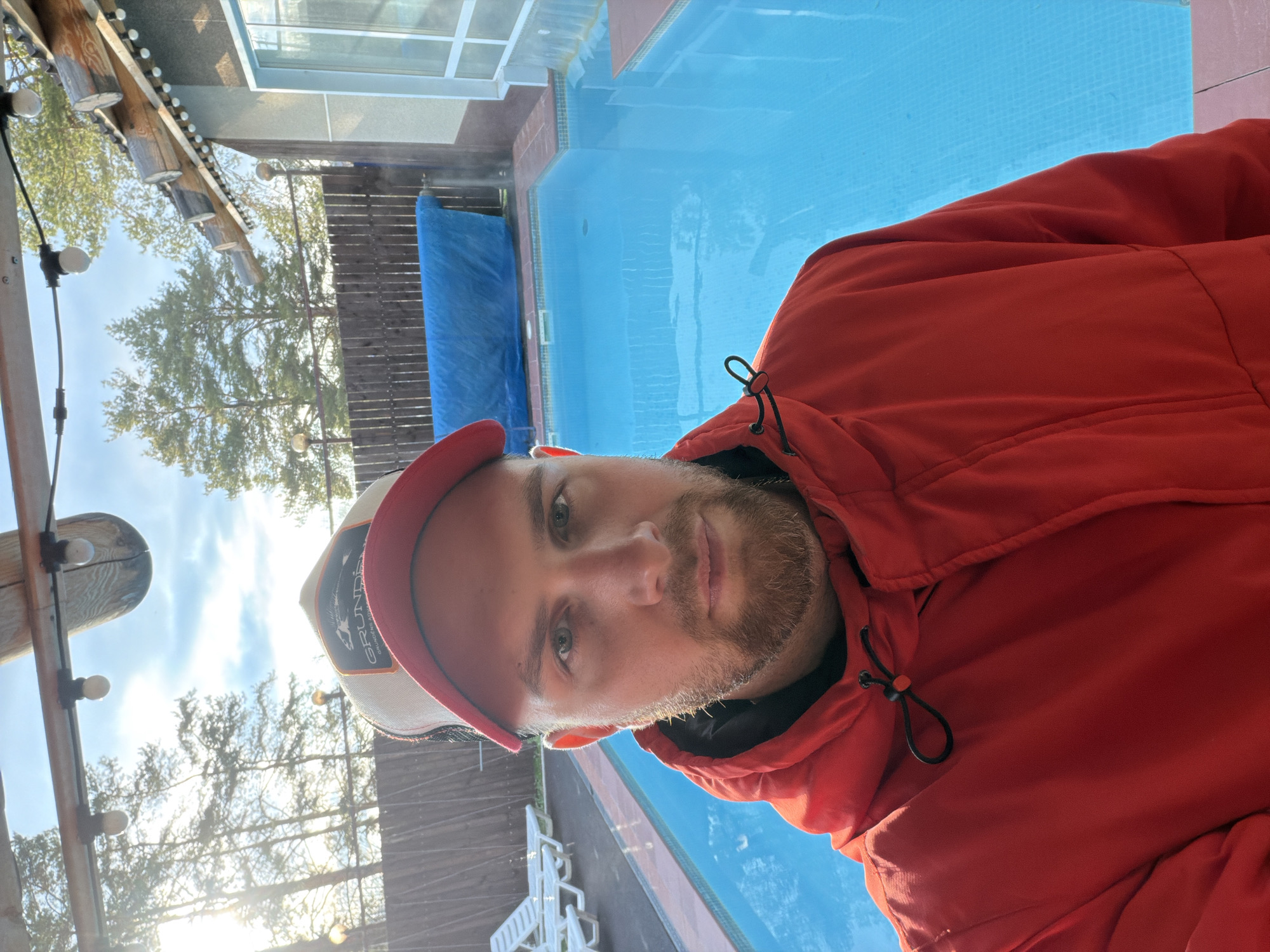 A man with a beard and mustache, wearing a bright red hooded jacket and a baseball cap with a red brim, takes a selfie outdoors on a sunny day in Chaikovsky, Russia. The cap has a logo partially visible, reading "GRUNDÉNS". The man looks directly at the camera with a neutral expression.
Behind him, a bright blue swimming pool with light blue tiles is a prominent feature. To the left, a blue tarp or cover is seen beside a wooden fence. In the background, green trees with visible foliage stand against a clear, sunny sky, with bright sunlight and some lens flare suggesting either morning or late afternoon. A wooden structure, possibly a pergola or roof support, equipped with string lights, adds to the outdoor ambiance. Several white lounge chairs are also visible near the pool area, indicating a leisure setting.