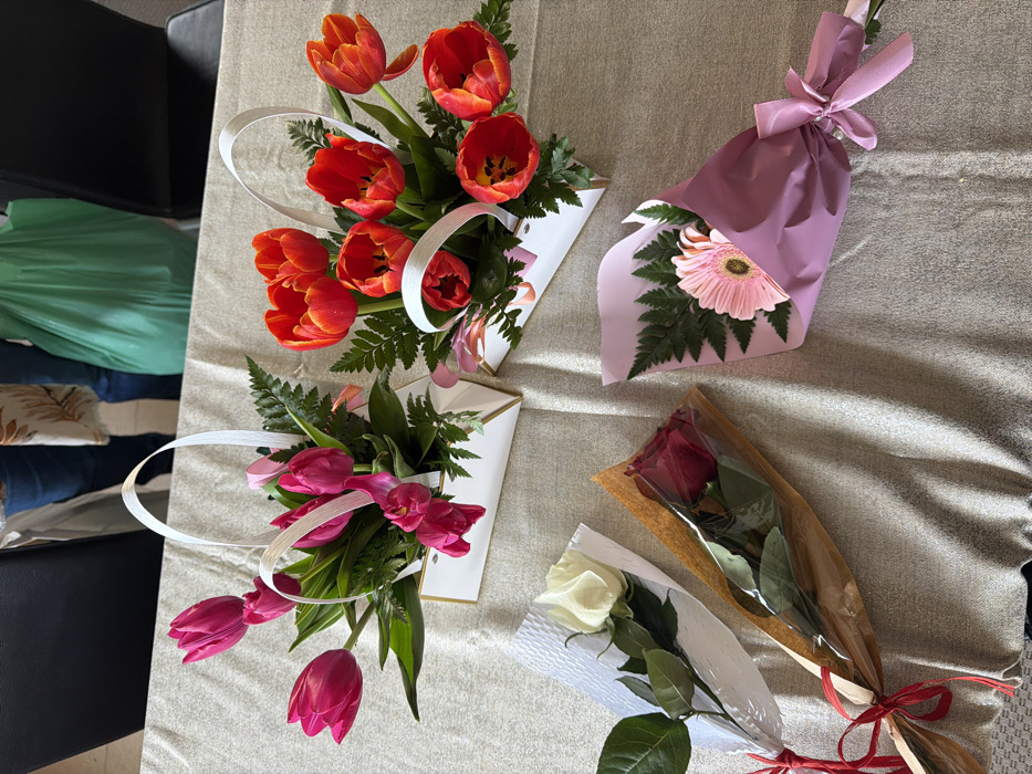 The scene is set indoors on a beige, textured tablecloth in the city of Vila Real, Portugal. Five distinct floral arrangements are displayed. Two are in white, rectangular basket-like containers with white ribbon handles. The left container holds multiple vibrant pink tulips interspersed with green fern-like foliage. The right container holds multiple bright red-orange tulips also interspersed with green fern-like foliage. Three individually wrapped single-stem bouquets lie below these arrangements. From left to right: a white rose with green leaves wrapped in white embossed paper and tied with a red ribbon; a dark red rose with green leaves wrapped in brown paper and clear plastic, secured with a red ribbon; and a pink gerbera daisy with green fern sprigs wrapped in light purple paper, tied with a matching purple ribbon. In the background, blurred figures of two individuals are partially visible, one wearing blue jeans and a light-colored patterned top, and a green plastic bag is also visible. Dark chair backs are subtly present.
