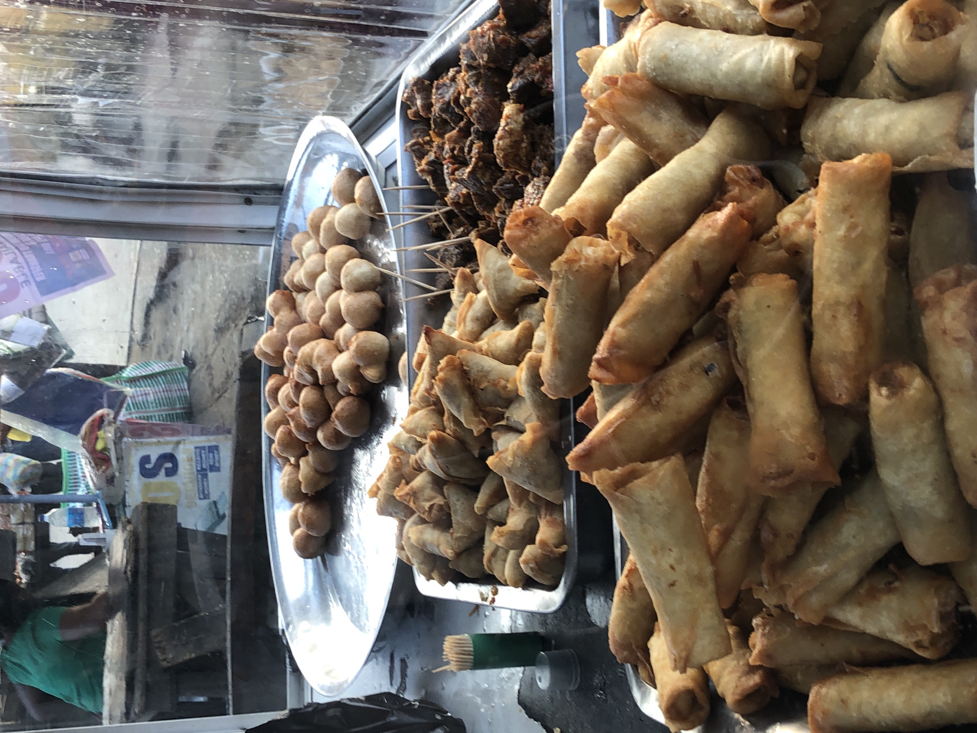 The image shows a street food stall in Ikeja, Nigeria, with various fried snacks displayed in metal trays. In the foreground, there are several trays filled with golden-brown spring rolls and what appear to be samosas. Behind these, a tray holds a pile of small, round fried dough balls. Further back, a tray contains pieces of fried meat. A person, partially visible in the background, is wearing a green shirt and appears to be preparing or serving food. The stall has a transparent covering, likely for protection from the elements, through which the scene is viewed. Signs and posters are visible behind the stall, and a water bottle is on a surface to the left. The overall setting suggests an outdoor market or roadside food vendor.