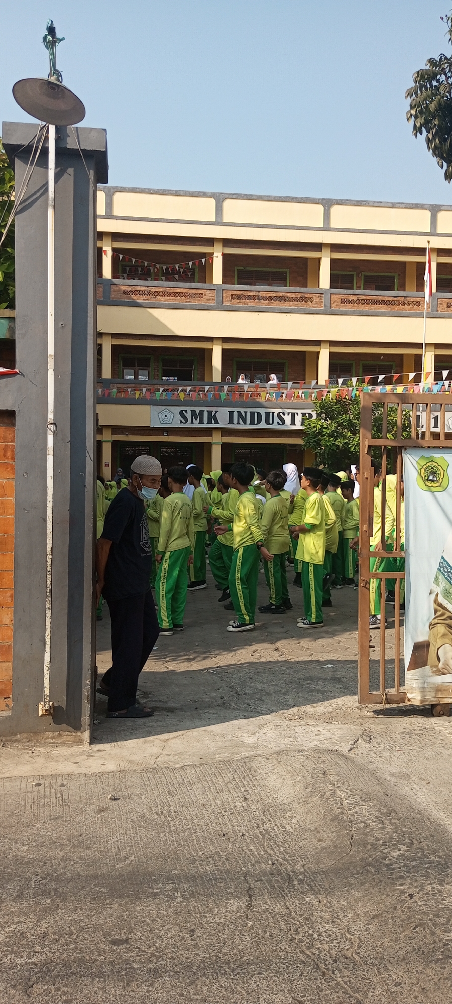 The image shows the entrance to a school building labeled "SMK Industri" in Setu, Indonesia. A group of students wearing bright green uniforms are gathered outside the building, appearing to be in formation or waiting for instruction. An adult male, wearing a dark shirt, dark pants, a white cap, and a face mask, stands near the entrance gate. The school building is a multi-story structure with a yellow facade and brick accents. Bunting and a flag are visible on the building's exterior, suggesting a possible event or celebration. The ground is paved with concrete. The sky is clear and blue, indicating a sunny day.