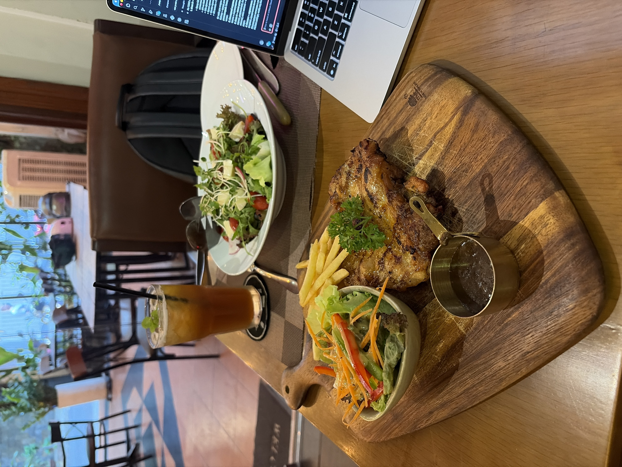 The image depicts a close-up view of a meal and a laptop on a wooden table in what appears to be a cafe or restaurant. In the foreground, a wooden board holds a grilled chicken steak, a small bowl of salad with carrots and lettuce, and a serving of french fries. A small metal cup contains a dark sauce. To the left of the meal, a tall glass filled with iced tea and a sprig of mint sits on a coaster. In the background, a white bowl of salad is visible, along with a laptop displaying code on its screen. A black backpack rests on a chair behind the laptop. The overall setting suggests a casual dining environment, possibly in Thủ Đức, Vietnam, given the presence of a laptop indicating work or study alongside a meal. The lighting is warm and inviting, with natural light streaming in from a window.