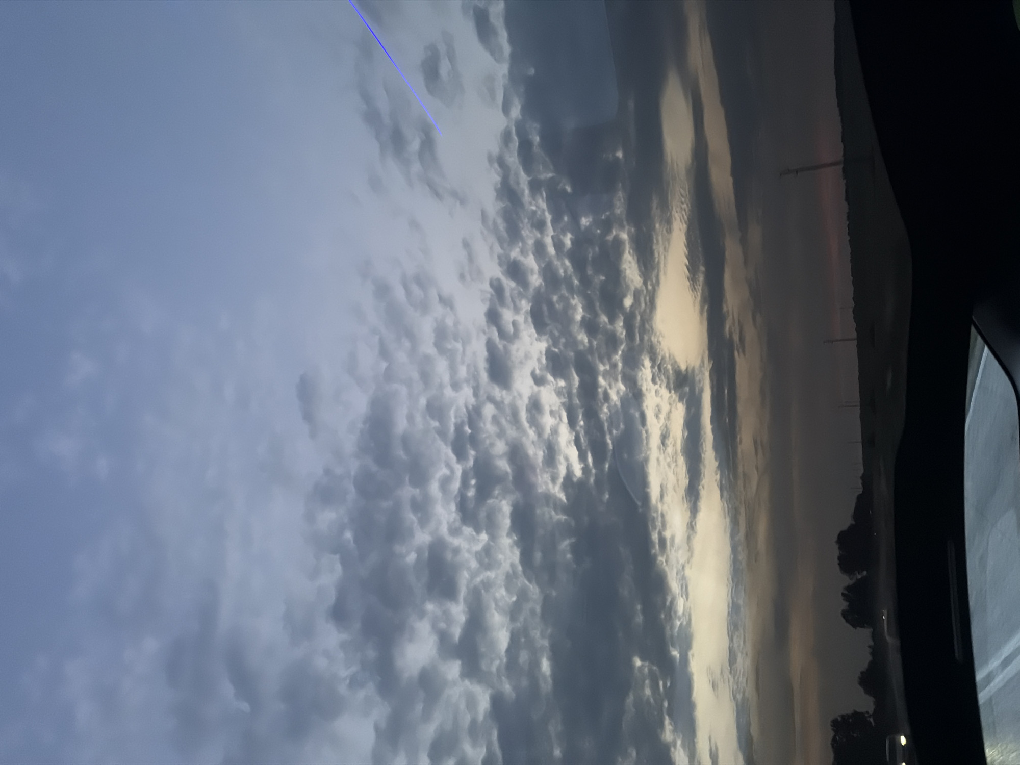 The image captures a dusk or dawn sky over a rural landscape, observed from the interior of a vehicle in Yasen, Bulgaria. The upper third of the frame shows a clear, muted blue sky transitioning downwards into a dense expanse of grey, lumpy altocumulus or stratocumulus clouds. Near the horizon, a band of warm, pale yellow and orange light indicates the position of the setting or rising sun beneath the cloud layer. The lower portion of the image reveals a dark, indistinct horizon line of flat terrain, likely fields, with the silhouettes of distant trees and a few utility poles. Faint headlights from other vehicles are visible in the far left foreground, suggesting a road. A distinct, bright blue, linear light streak angles from the upper right corner towards the center-right, appearing as a lens flare or an internal reflection. The bottom section of the image shows the dark contour of the vehicle's dashboard and reflections on the windshield. No individuals are visible.