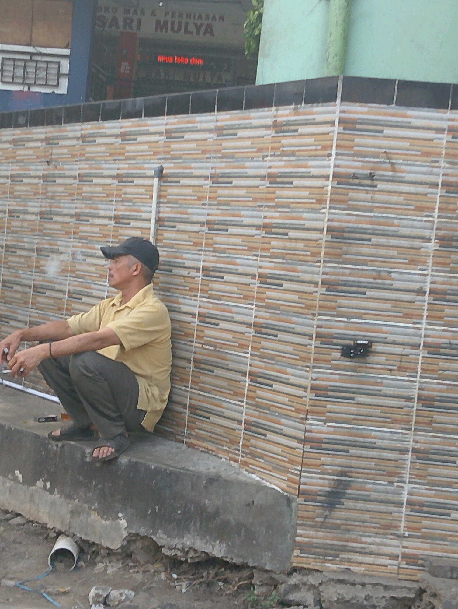 In Lembang, Indonesia, a man wearing a dark baseball cap and a tan polo shirt is squatting on a concrete ledge, leaning against a tiled wall. He is smoking a cigarette, with visible smoke rising from it. His left arm rests on his bent left leg, and his right arm is also bent, with his hand near his left thigh. He is wearing dark pants and dark sandals. The wall behind him is covered in horizontal rectangular tiles of varying colors, including beige, brown, white, and gray. To the left of the man, a white pipe protrudes from the wall. In the background, a blurred sign reads "OKO MAS & PERHIASAN SARI MULYA," indicating a shop. The ground in front of the ledge is rough and appears to be dirt with some debris, including a white pipe opening and loose stones.