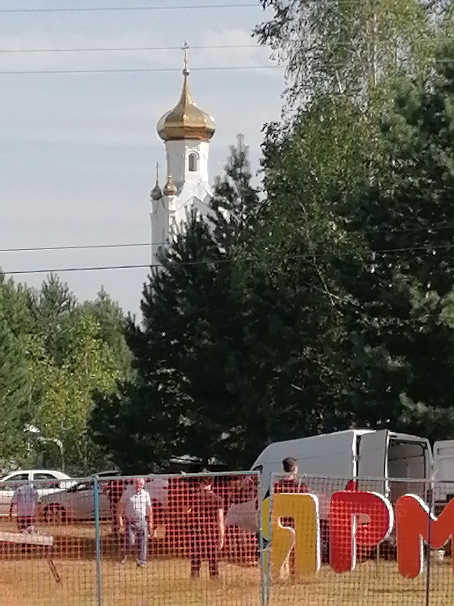 The image captures a scene in Shadrinsk, Russia, with a prominent white church featuring a golden dome dominating the background. In the foreground, a red and white checkered fence separates the viewer from a gathering of people. A white van with its doors open is visible behind the fence, along with large, colorful letters that appear to spell out part of a word or phrase, possibly related to an event or festival. Several individuals are present, some seemingly engaged in conversation or activity, while others stand near vehicles parked in the vicinity. The environment appears to be outdoors, with trees and foliage suggesting a rural or park-like setting within the city. The overall impression is that of an organized event or gathering taking place on a clear day.