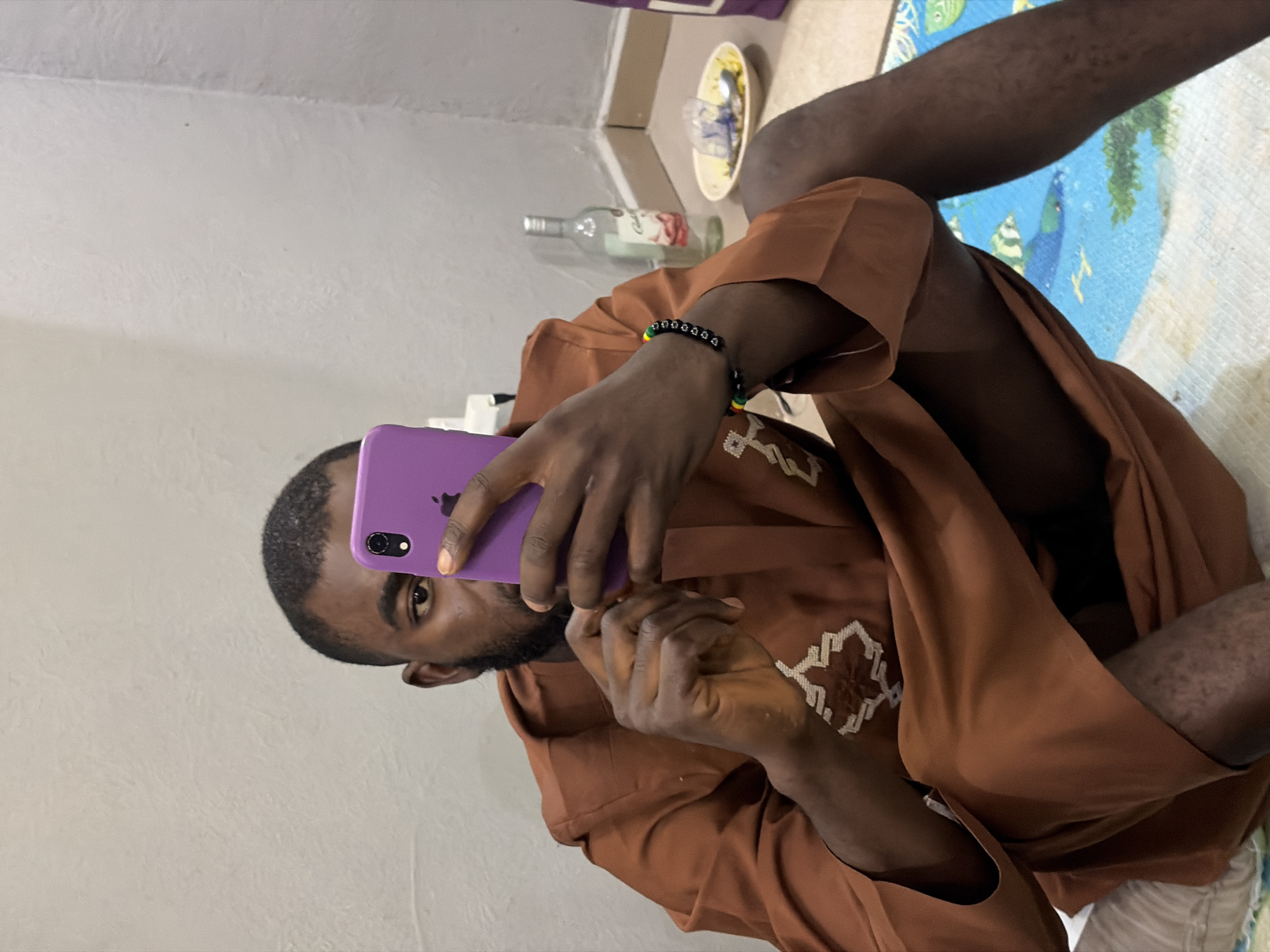 A person with dark skin is seated on the floor, holding a purple smartphone in their left hand and looking at the screen. They are wearing a brown robe with white embroidered patterns on the chest. A bracelet with black, green, yellow, and red beads is on their left wrist. Behind the person are a bottle, a plate with food, and a patterned mat on the floor. The background includes a plain white wall. The location is potentially a residence in Jos, Nigeria, based on the person's appearance and the environment's characteristics.