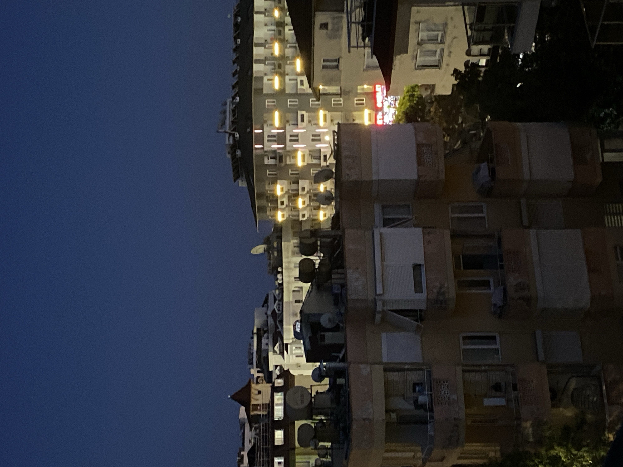 The image, originally rotated, captures an urban night scene in Alanya, Turkey. The frame is dominated by multi-story apartment buildings on the right, set against a deep blue, clear night sky on the left. The sky shows no clouds, suggesting calm weather.
Numerous windows in the buildings are lit, casting warm yellow and orange glows that stand out against the otherwise dimly lit structures. The facades of these buildings are densely populated with various objects, including a multitude of air conditioning units attached below windows and several satellite dishes. Balconies and other architectural details are visible on the structures.
Between some of the buildings and at their bases, dark green foliage from trees can be discerned. On one of the more central buildings, a prominent red neon sign is visible. The text appears to spell out "HOTEL," suggesting a commercial establishment amidst the residential blocks.
No people or explicit activities are visible in the scene, which portrays a quiet, illuminated urban landscape during the nighttime hours.