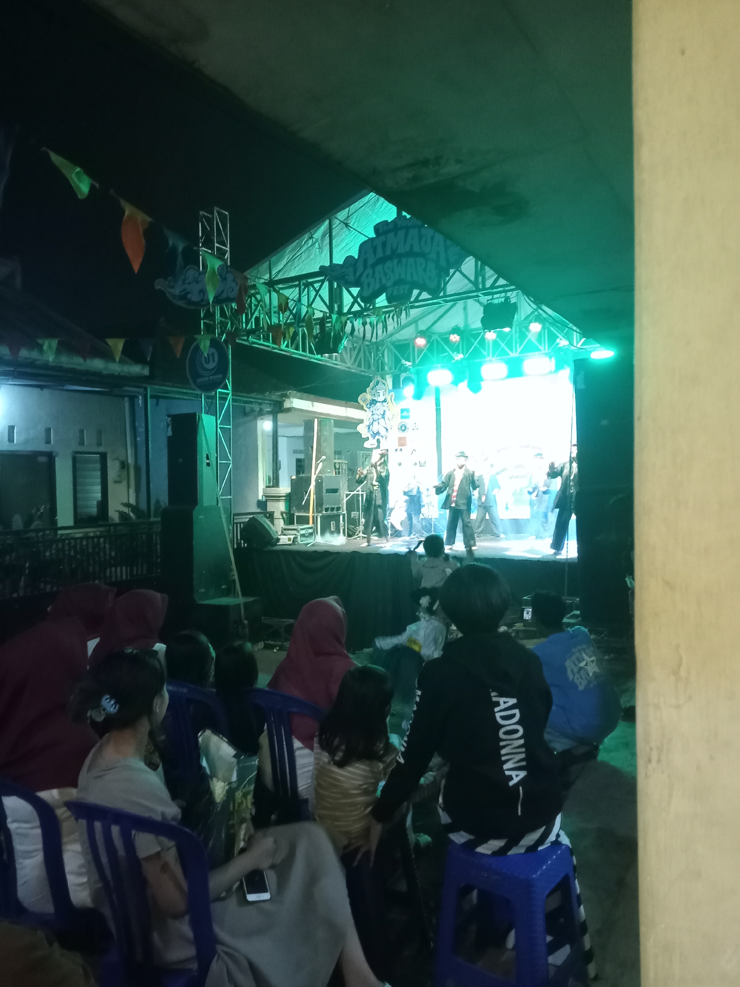 The image depicts an outdoor stage performance at night, likely in the city of Batu, Indonesia, indicated by cultural elements and the general ambiance. The stage is illuminated by bright green and white lights, with a banner above reading "ATMADJA BASHARA FEST". Several performers are visible on stage, some in traditional attire, engaging in what appears to be a cultural dance or performance. In the foreground, a crowd of spectators, including adults and children, are seated on plastic chairs, observing the event. The environment is dark, typical of an evening outdoor setting, with some surrounding buildings and a wall visible. Decorative flags are strung across the top of the scene, adding to the festive atmosphere. The overall impression is one of community gathering for a cultural celebration.