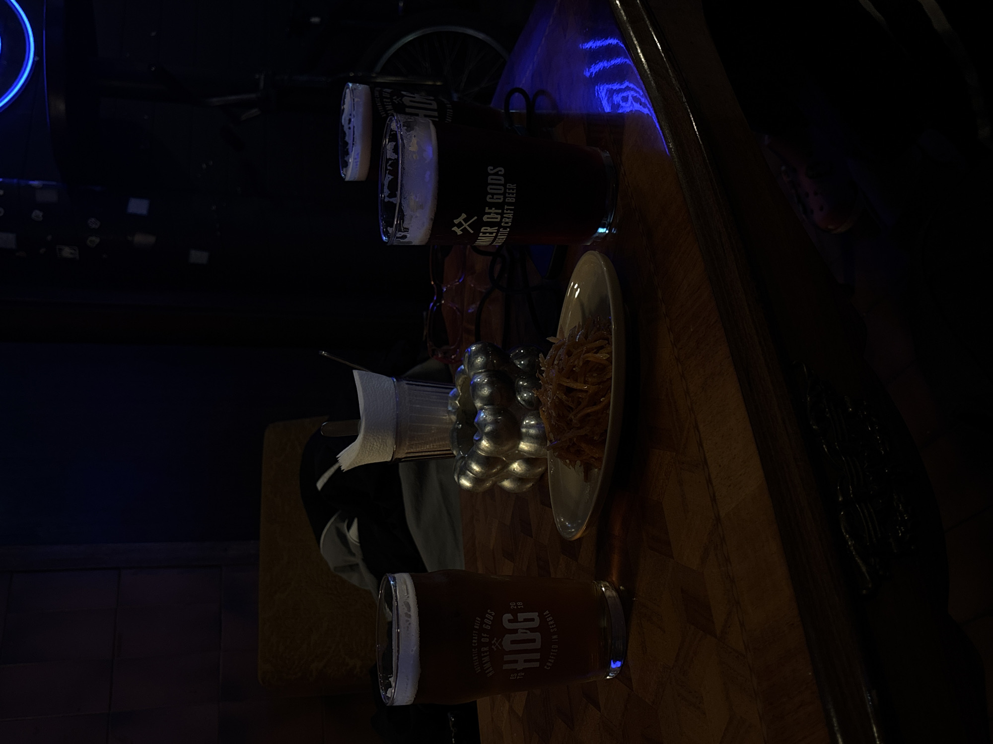 This image captures a scene at a bar in Belgrade, Serbia. On the bar surface, there are three beer glasses, each filled with a dark brew. The brand name "Hammer of Gods Authentic Craft Beer" and "HOG Crafted in Serbia" can be clearly identified on the glasses. Adjacent to the beer glasses, there is a plate filled with what appears to be fried food, possibly fries or some other savory snack. A metallic centerpiece is also visible on the counter. In the background, a person is seated. The setting is dimly lit, and a blue neon light casts a glow on parts of the scene, adding to the bar's ambiance.