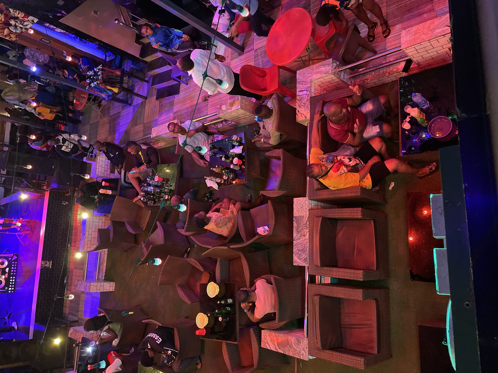 The image depicts a top-down view of an outdoor lounge, possibly in Obosi, Nigeria, with multiple seating areas comprised of wicker chairs and tables. Patrons are scattered throughout, engaging in conversations and leisure activities. The lighting is a mix of ambient and decorative string lights, creating a vibrant atmosphere. A bar area is visible along one edge, and a stage with audio equipment indicates a possible entertainment venue. The scene suggests a relaxed, social environment.