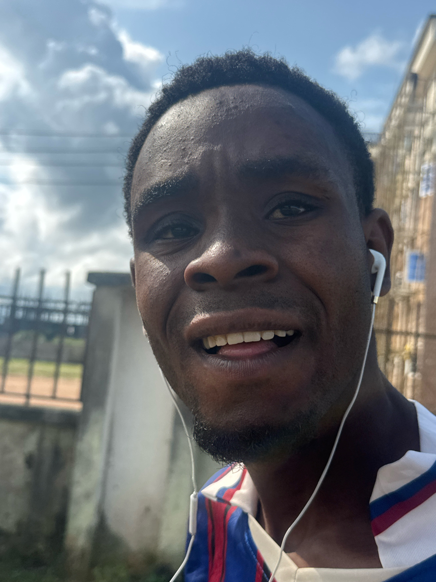 A close-up selfie of a dark-skinned man wearing white earbuds is captured outdoors. He has dark, curly hair and a faint beard. His mouth is slightly open, revealing his teeth, and he appears to be looking directly at the camera with a neutral expression. He is wearing a white shirt with red and blue stripes. In the background, a blurred fence can be seen on the left, and a building with windows is visible on the right. The sky above is partly cloudy. The setting is likely the city of Ihiagwa, Nigeria, given the context.