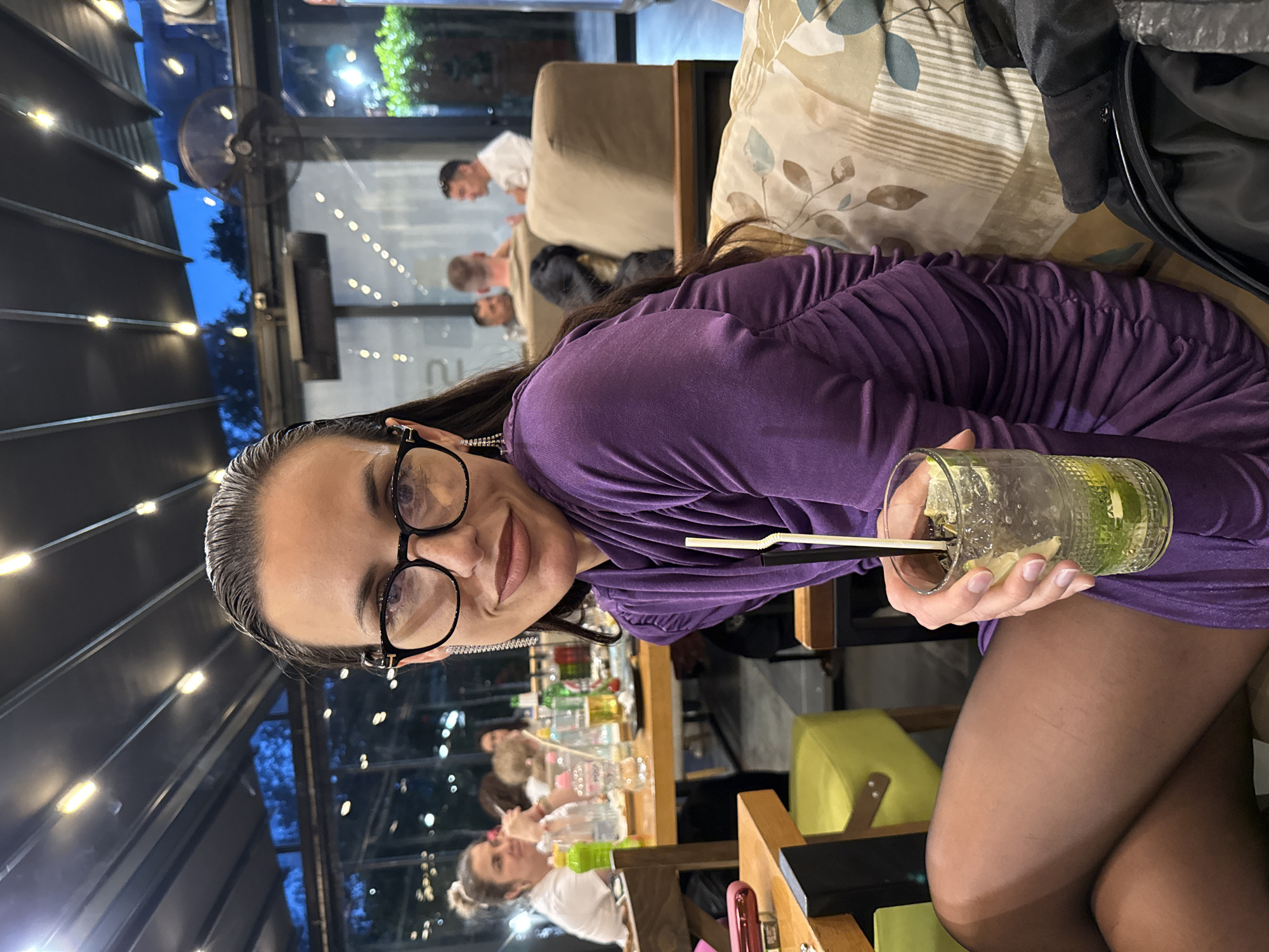 A woman with dark hair pulled back, wearing glasses and a purple dress, holds a cocktail in a clear glass with a straw. She appears to be seated at a table, with other people visible in the background and in the reflection of a glass partition. The setting appears to be a restaurant or bar, as suggested by the tables, chairs, and bar equipment in the background. The location is Pleven, Bulgaria.