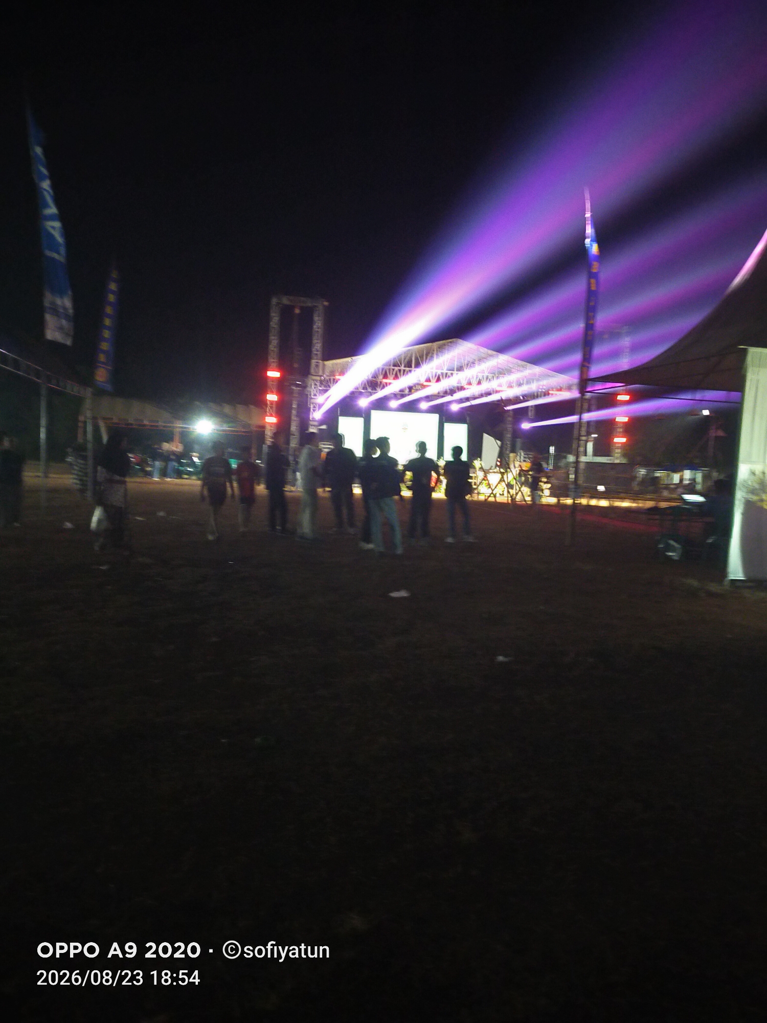 The image captures a nighttime outdoor event, likely a concert or festival, illuminated by vibrant purple and white stage lights. In the background, a large stage structure with a white tent roof is visible, adorned with numerous spotlights casting beams into the dark sky. Several people, mostly silhouetted, are scattered across the muddy ground in front of the stage, some appearing to watch the performance. Two flags, one blue and white, the other yellow and blue, are partially visible on the left side, suggesting sponsorship or team affiliation. The ground is covered with dark soil and appears uneven, consistent with an outdoor venue in a rural area. The timestamp and camera information "OPPO A9 2020 - ©sofiyatun 2026/08/23 18:54" are overlaid at the bottom left of the image, indicating the date and time the photo was taken. The location is identified as Cluwak, Indonesia.