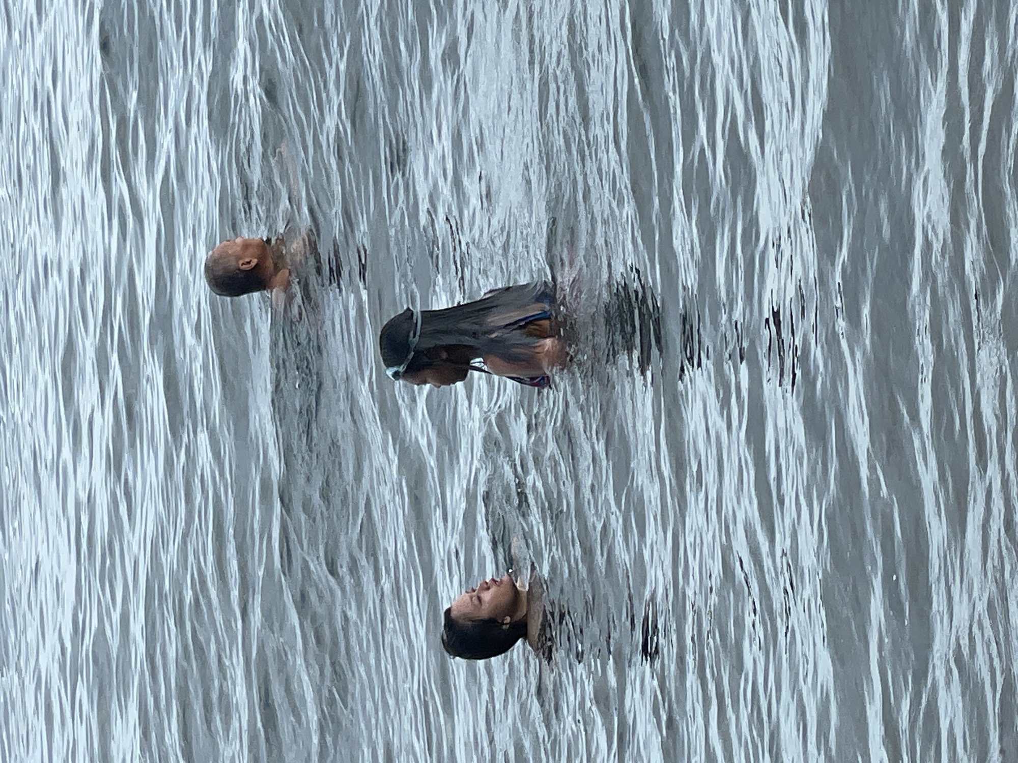 In the calm, rippling waters off Dauin, Philippines, three individuals are visible. In the background, a person with a shaved head is swimming, facing to the right. In the mid-ground, a person with dark hair and swimming goggles on their head is in the water, facing left and engaged in conversation with another person. This second person, in the mid-ground to the left, has dark hair and is also swimming, looking towards the person with goggles. The environment is an open body of water with moderate waves.
