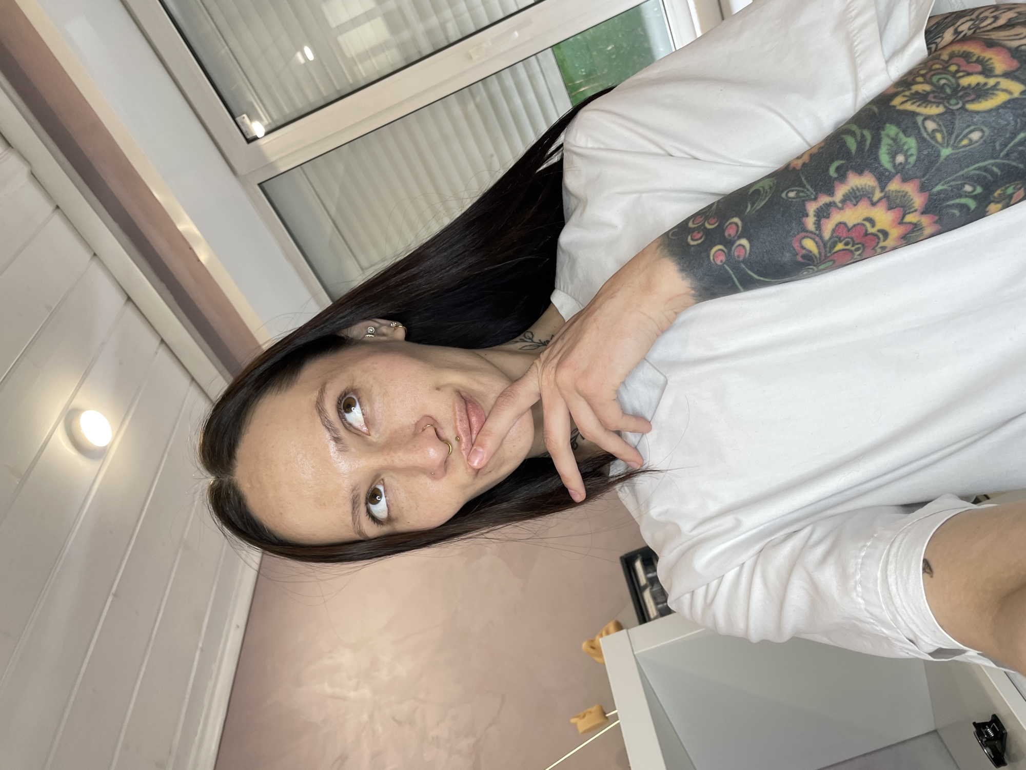 A fair-skinned woman with dark brown hair is shown from a high-angle view, wearing a white t-shirt. She has tattoos on her left arm, visible above the elbow. Her left hand is near her mouth, and she is looking up. She has pierced ears and a pierced nose. The background includes a window with blinds and a light pink wall. A portion of a white cabinet is visible in the lower right corner. The location in Izhevsk, Russia is not discernible from the image.