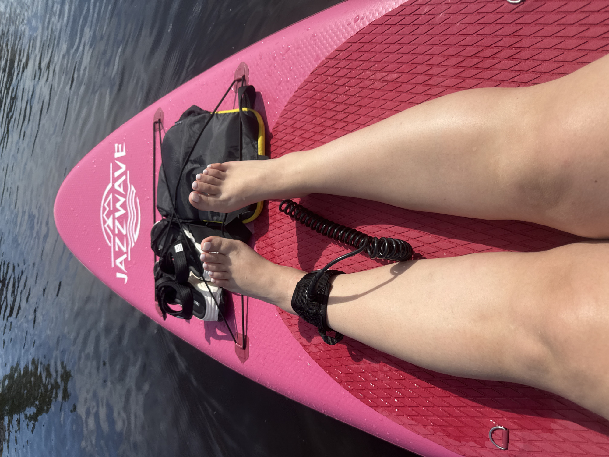 The scene is situated on water, presumed to be in the city of Vologda, Russia as per context. A first-person perspective captures the lower body of an individual, specifically their legs and feet, resting on a pink paddleboard. The left leg is bent with the foot positioned towards the board's bow, wearing a black ankle leash with a coiled black cord extending forward to an attachment point on the board. The right leg is extended forward. Both feet display light-colored nail polish. Ahead of the feet, a pair of black and white platform sandals and a black waterproof dry bag with yellow accents are secured on the paddleboard. The paddleboard, branded "JAZZWAVE" in white lettering near its bow, features a textured grip pad in a darker pink hue, with water droplets visible on its surface. The surrounding water is dark with slight ripples, reflecting ambient light. The scene is brightly lit by natural sunlight, casting distinct shadows from the legs and objects onto the paddleboard surface. A metal D-ring is visible on the port side of the paddleboard.