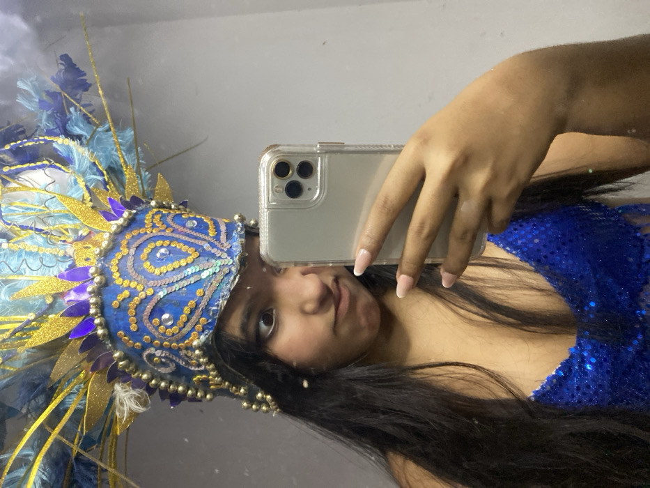 A person wearing a blue, embellished headdress with feathers and sequins takes a selfie in a mirror with an iPhone in a clear case. She has long dark hair and wears a blue sequined top. Her skin tone is tan, and she has manicured nails. The background is a plain, light-colored wall. The photo is taken at an angle, presenting a slightly distorted view of the scene.