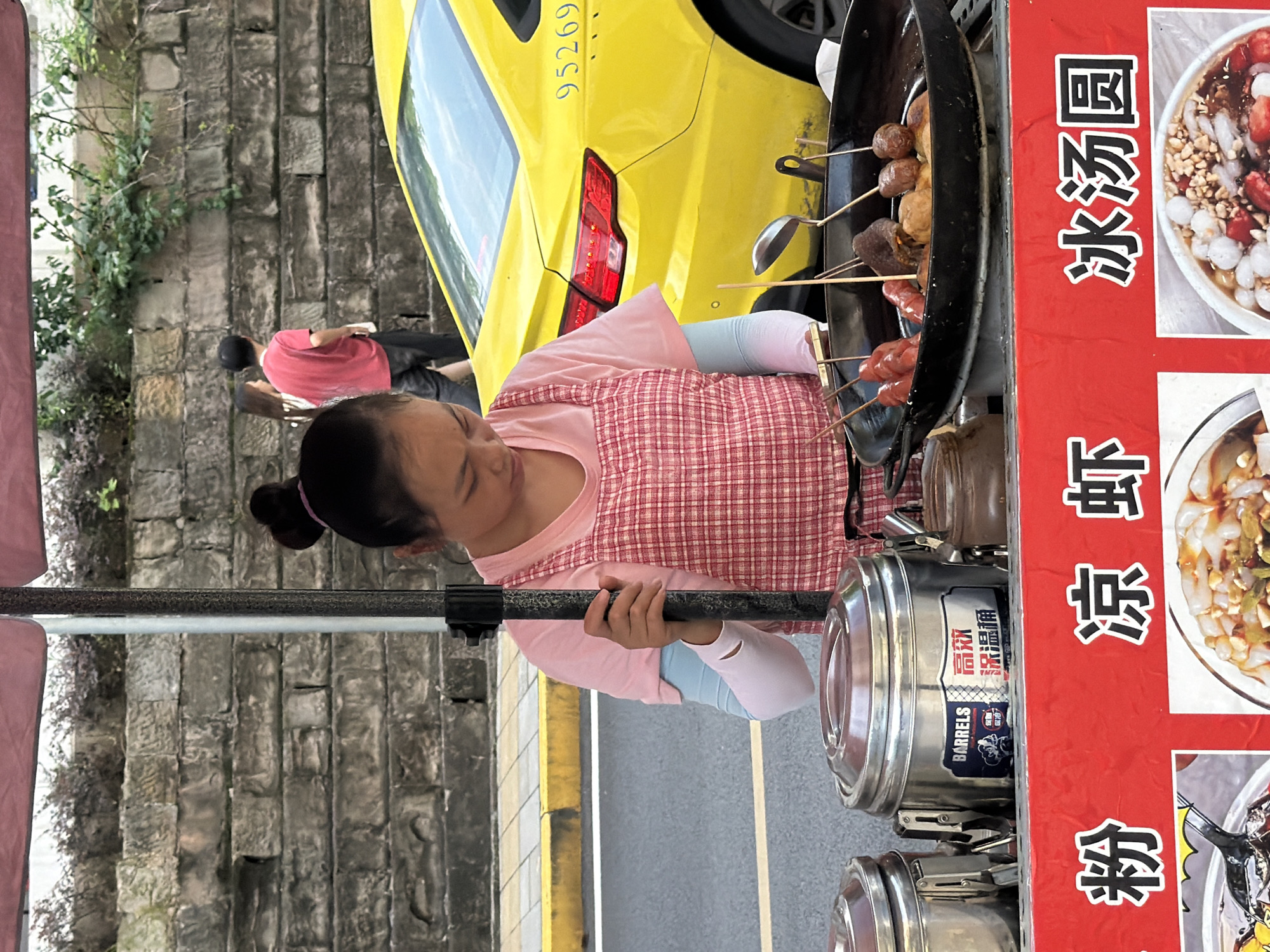 A street vendor, a woman with her hair in a bun, stands behind a food cart under a pink umbrella. She is wearing a pink t-shirt and a red and white checkered apron. In front of her is a large black wok filled with fried food items on skewers, including sausages and meatballs. To her left are two silver food containers with lids, one labeled "BARRELS" and with Chinese characters. A yellow taxi with license plate "95269" is parked behind the food cart. In the background, a stone wall with some greenery and two people walking are visible. The foreground features a red banner with Chinese characters, possibly indicating food items for sale. The scene is located on a city street in Yuzhong District, China.